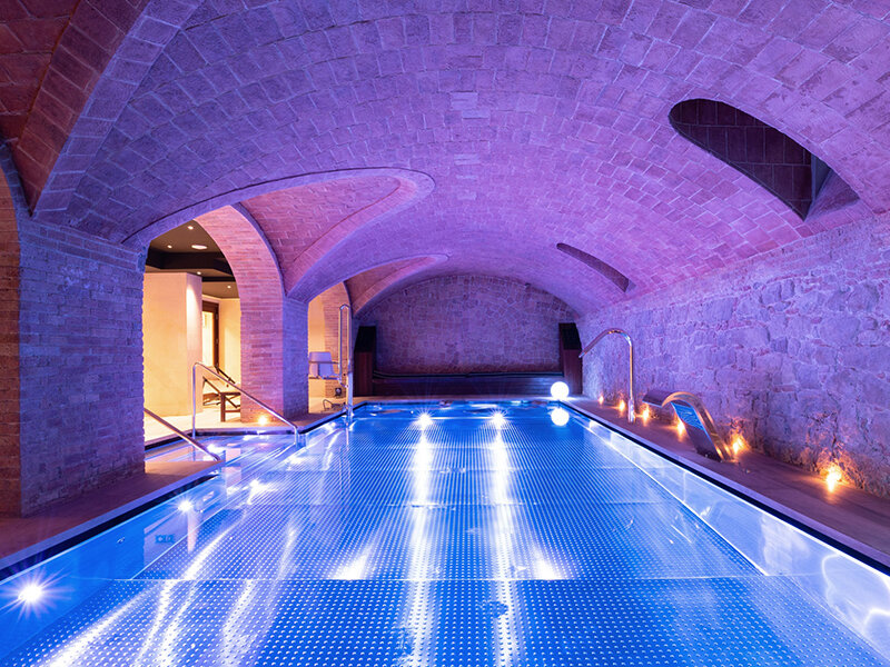 Piscina interior con agua iluminada de azul, techo abovedado de ladrillo y elementos de spa.