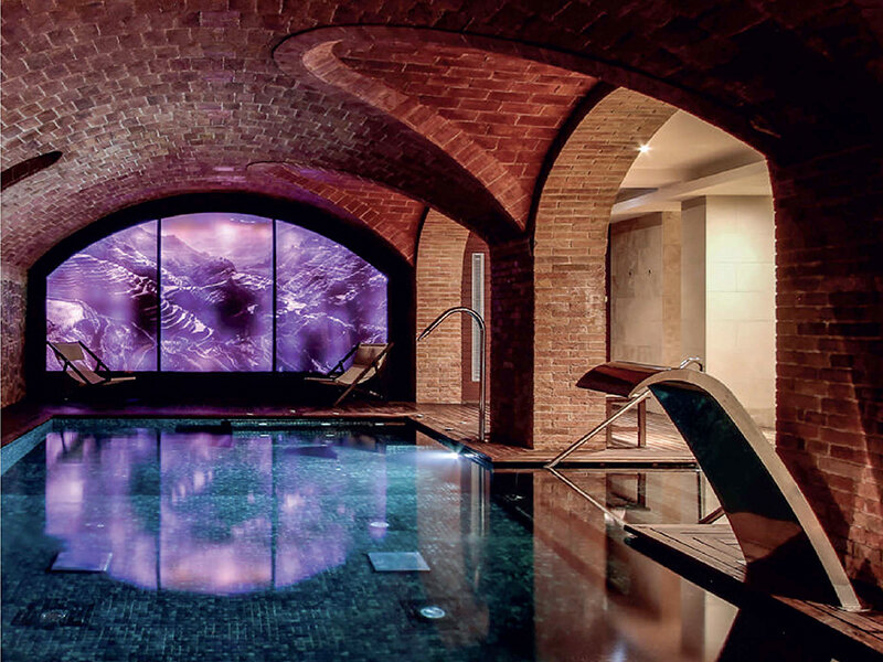 Piscina interior con techo abovedado de ladrillo y una gran pantalla digital con paisaje montañoso.