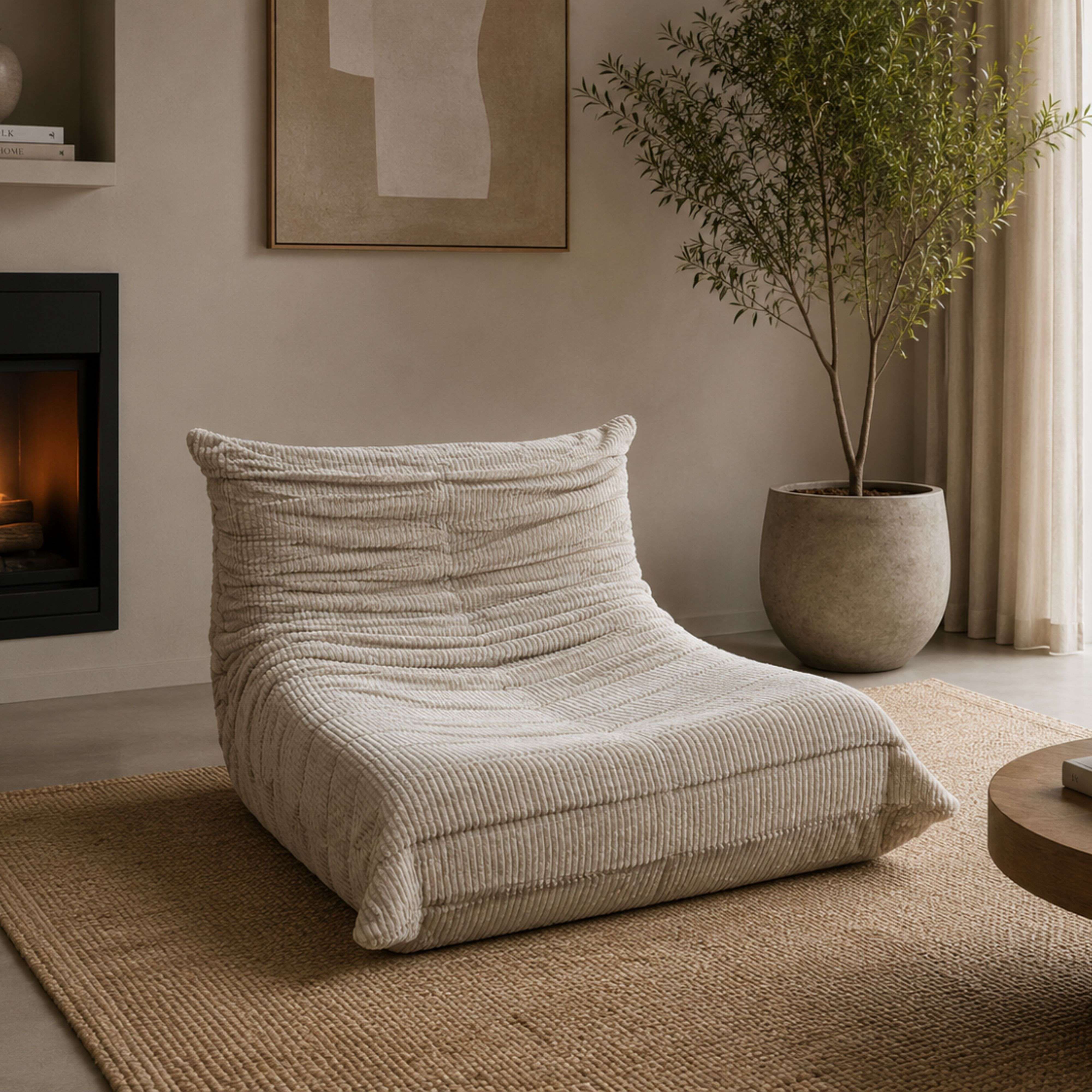 Un sillón beige de pana descansa sobre una alfombra de yute, junto a una chimenea y una planta.