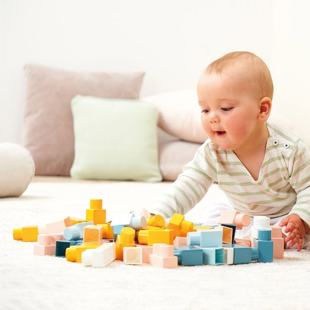 Un bambino gioca con blocchi di legno colorati su un tappeto, cuscini sullo sfondo.