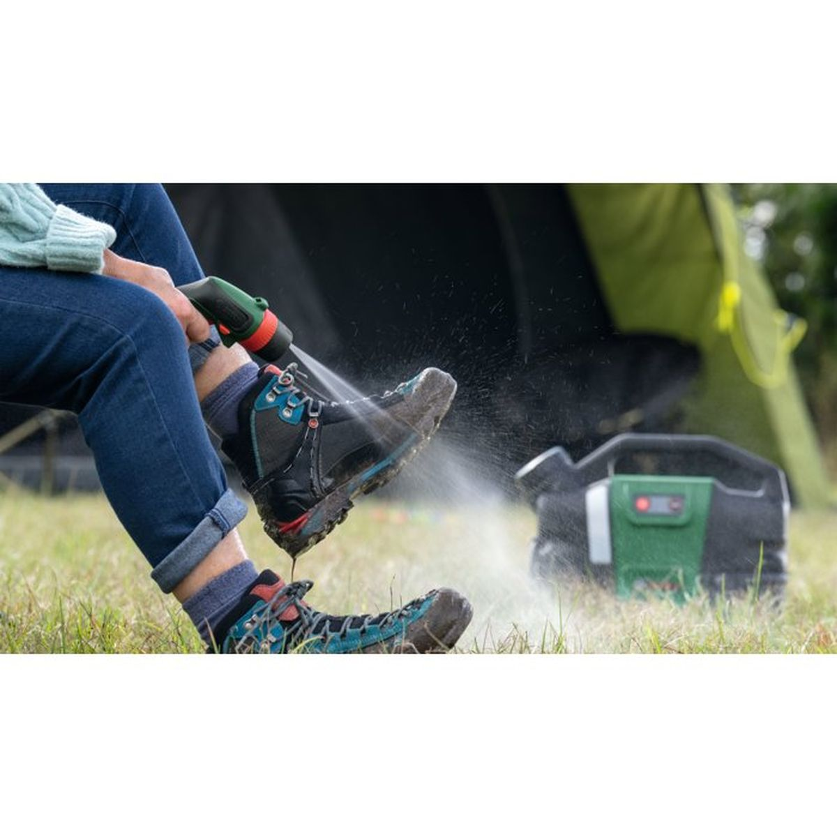Eine Person wäscht Wanderschuhe mit einem Schlauch neben einem Zelt und einem Aggregat.