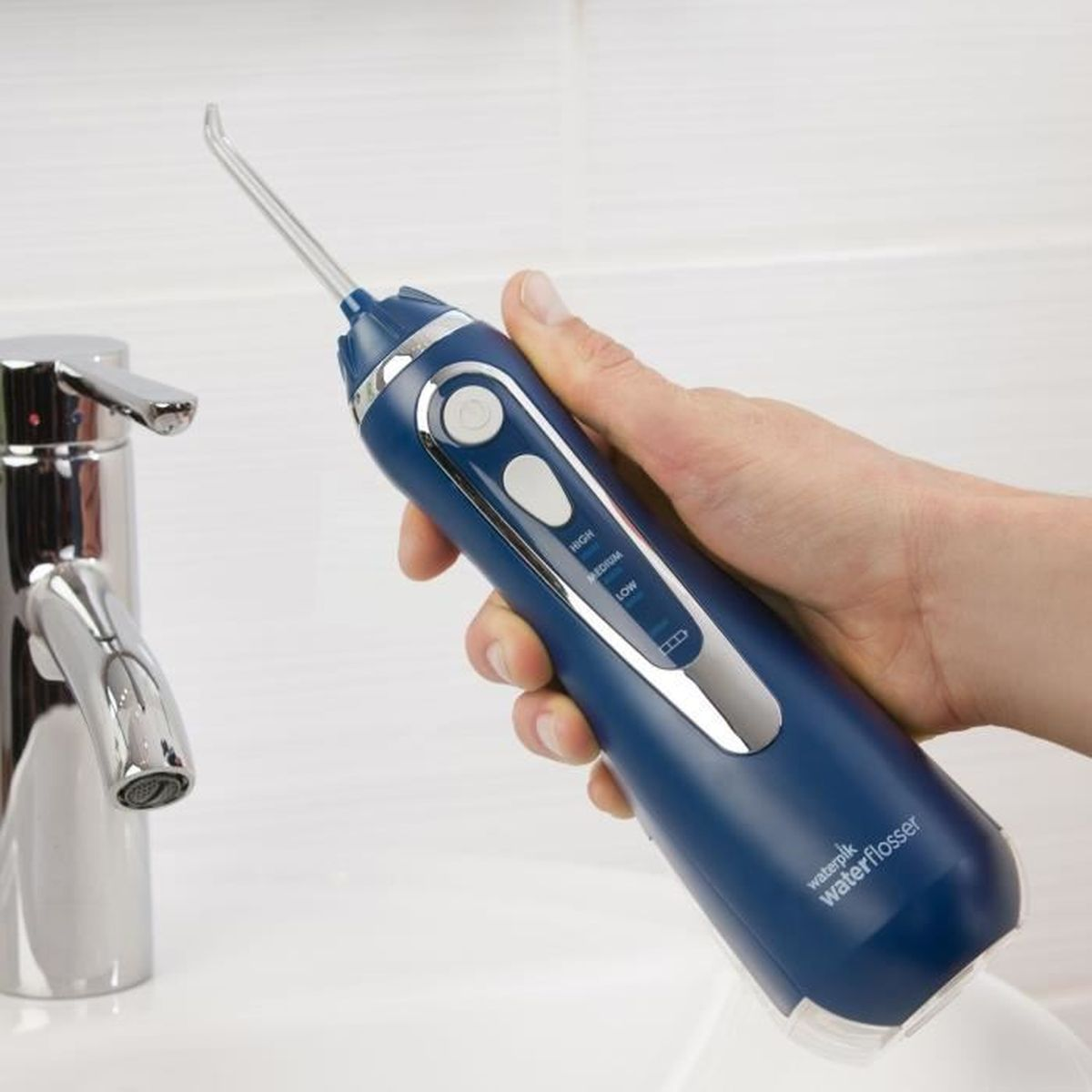 Eine Hand hält eine blaue Waterpik-Zahnseide neben einem Wasserhahn im Waschbecken.