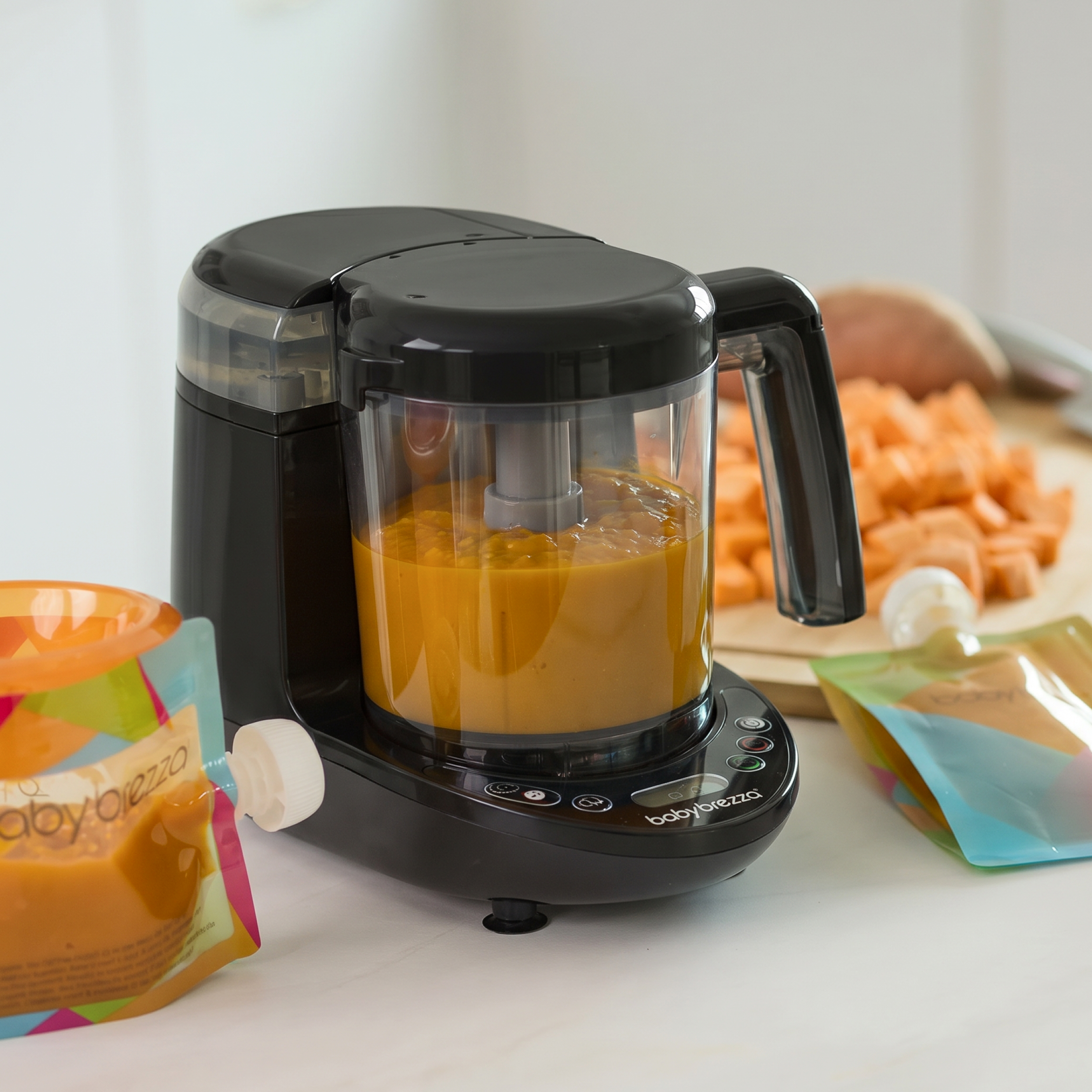 Ein schwarzer Babybrei-Zubereiter mit orangefarbenem Brei, daneben Quetschbeutel.