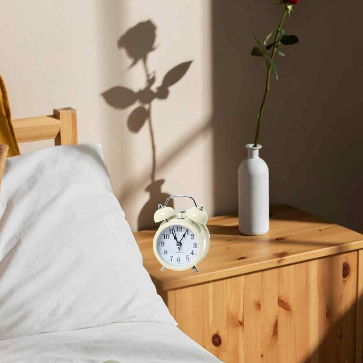 Ein cremefarbener Wecker steht auf einem Nachttisch aus Holz neben einer weißen Vase mit einer Rose.