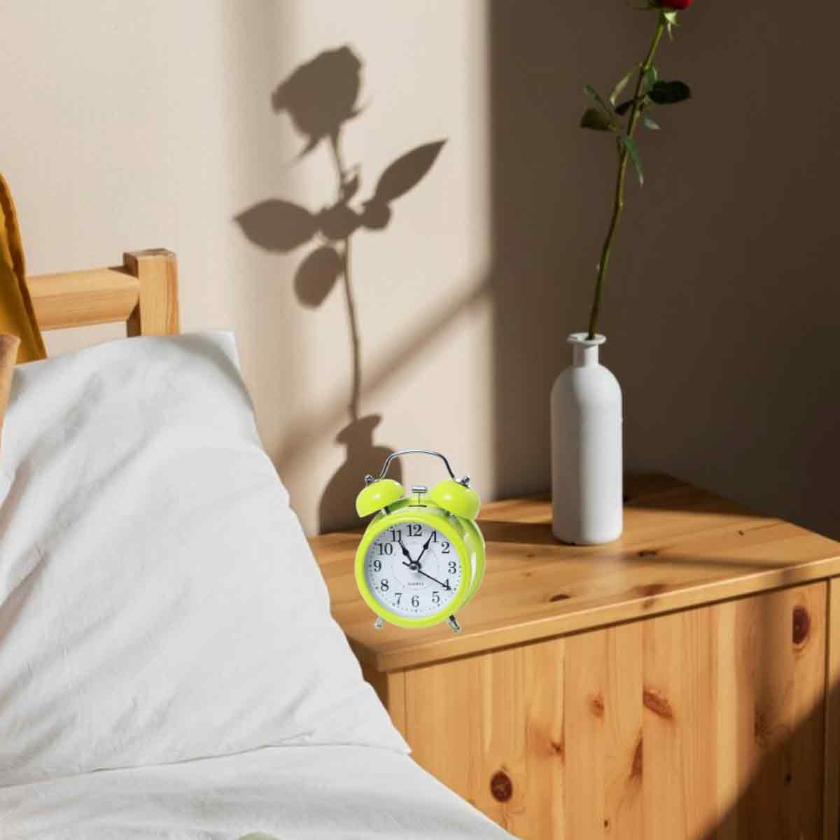 Ein grüner Wecker auf einem Nachttisch neben einem Bett. Eine Rose im Hintergrund.