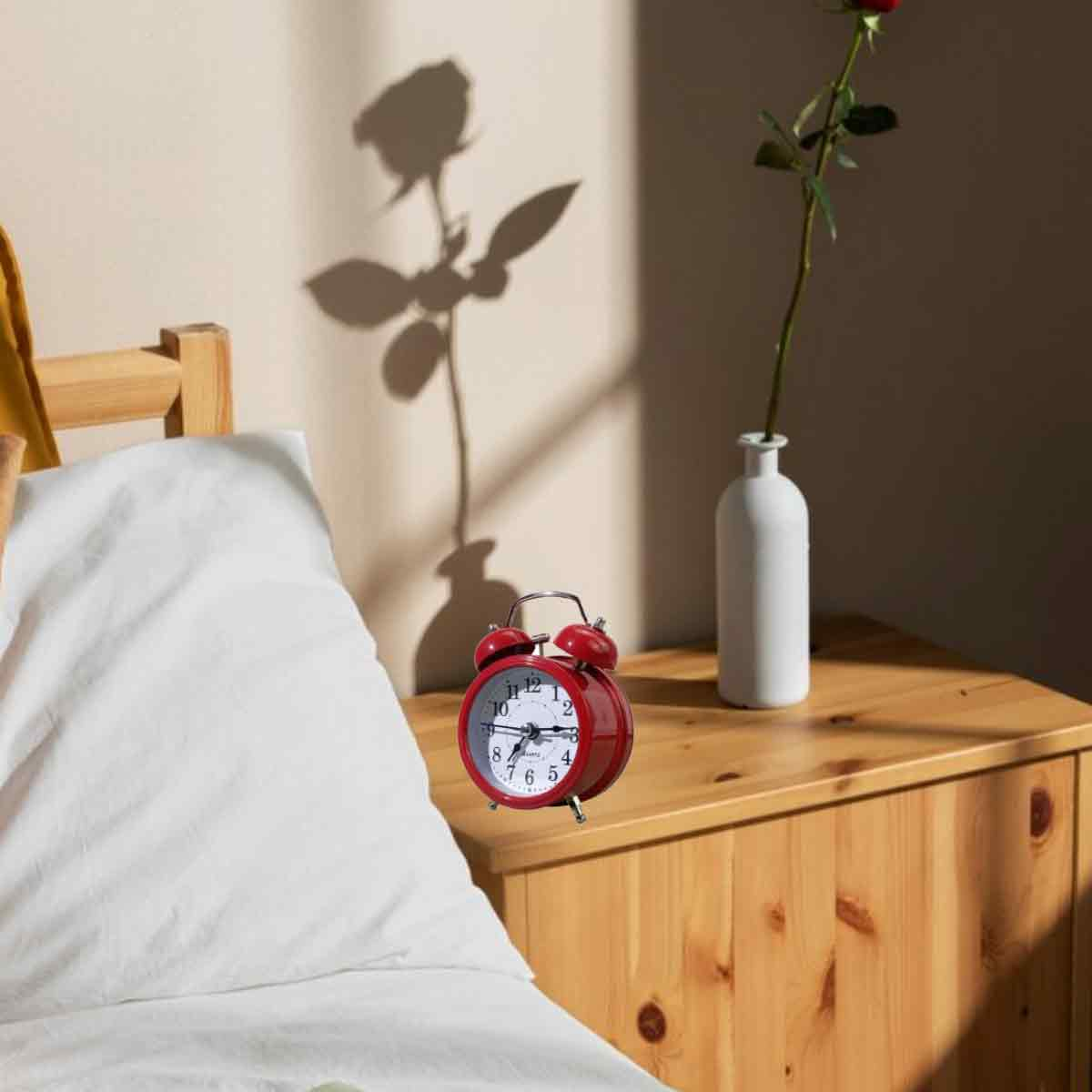 Ein roter Wecker und eine weiße Vase mit Rose auf einem Holztisch.