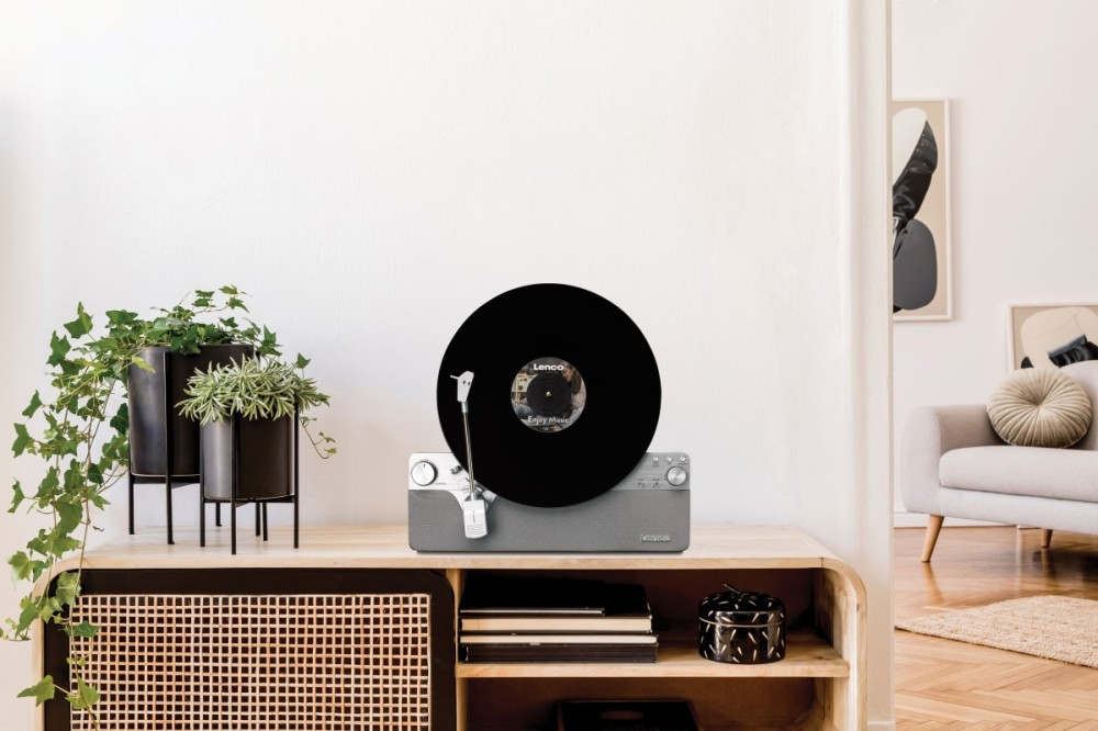 Gramofon z czarną płytą winylową stoi na drewnianej szafce. Po lewej dwie doniczki z roślinami.