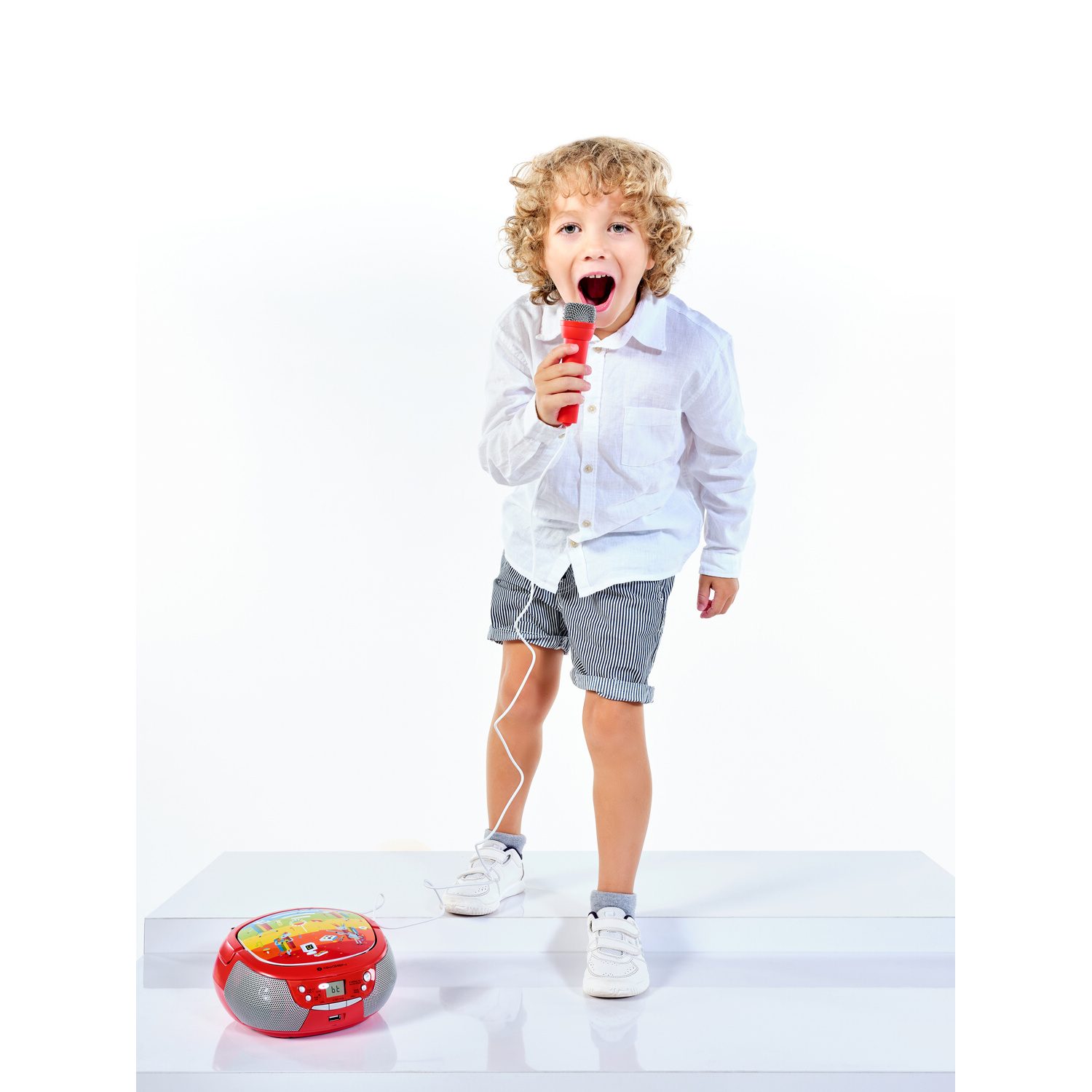 Un bambino con i capelli ricci canta in un microfono rosso, vicino a un lettore CD.