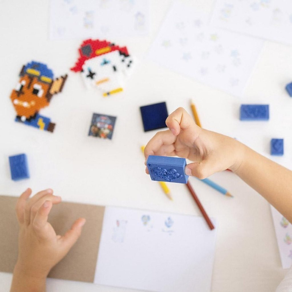 Un niño sostiene un sello azul con un patrón; lápices de colores y arte de cuentas esparcidos.