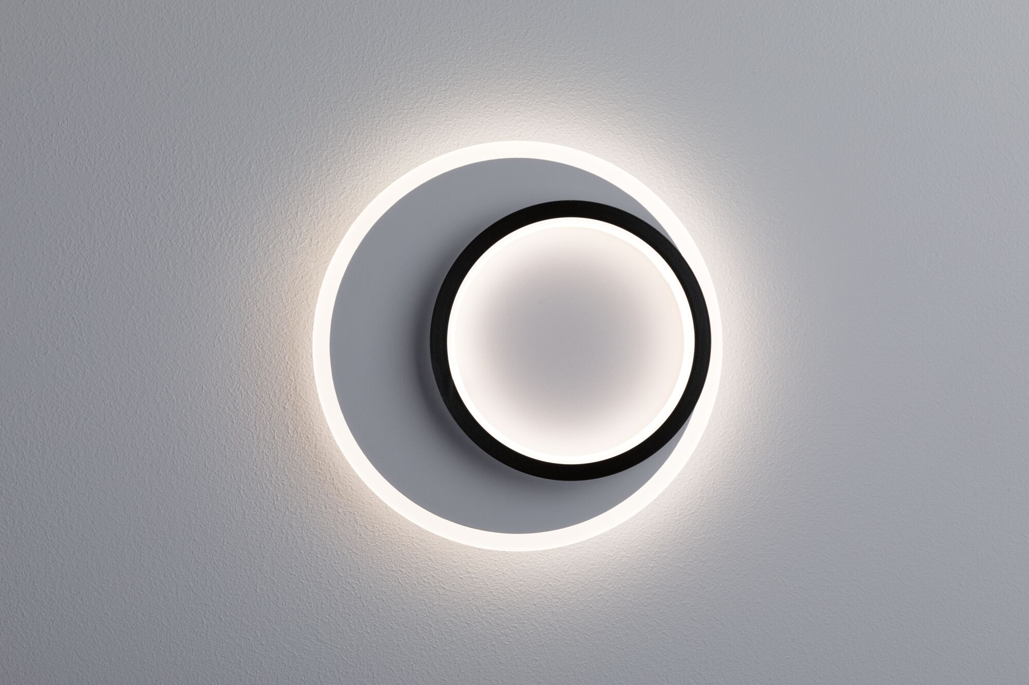 Eine runde Wandleuchte mit schwarzem Ring und weißem Licht auf grauem Grund.