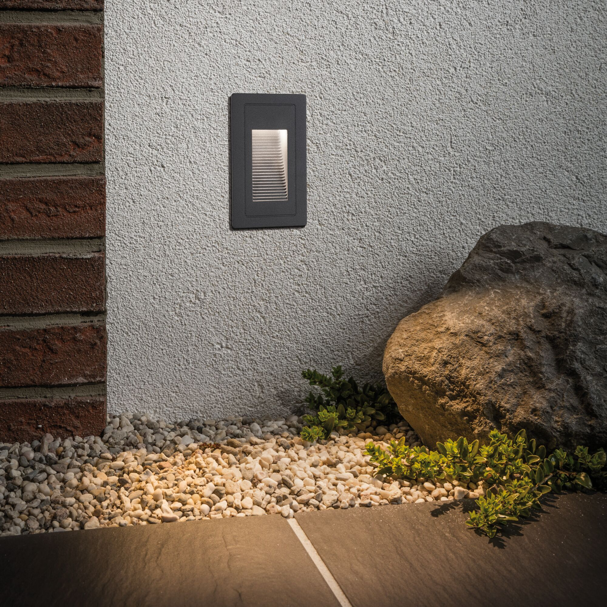 Una lampada da parete illumina ciottoli, piante e una grossa roccia vicino a un muro di mattoni.
