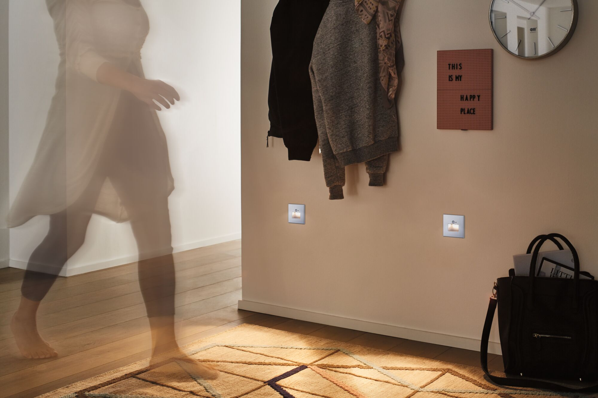 Eine Person geht durch einen Flur mit Teppich, Jacken an der Wand und einer Tasche.