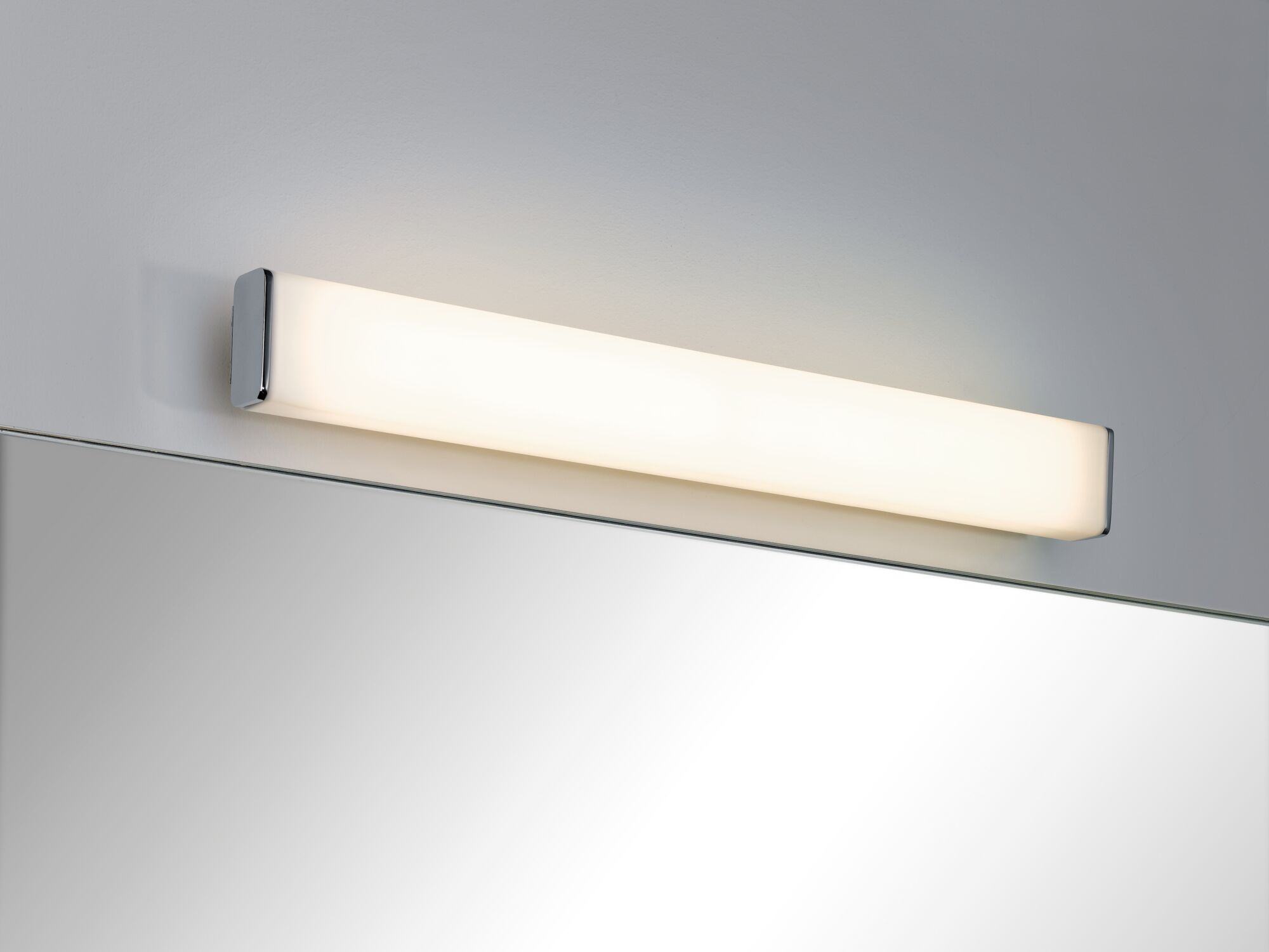 Una lampada da parete rettangolare con bordo cromato brilla sopra uno specchio.