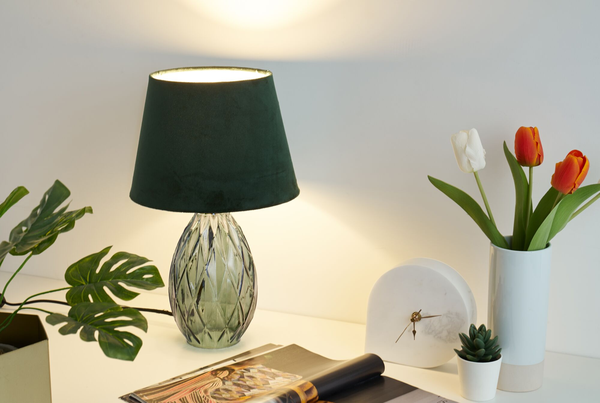 Eine grüne Lampe beleuchtet einen Schreibtisch mit Blumen, einer Uhr und einer Zeitschrift.