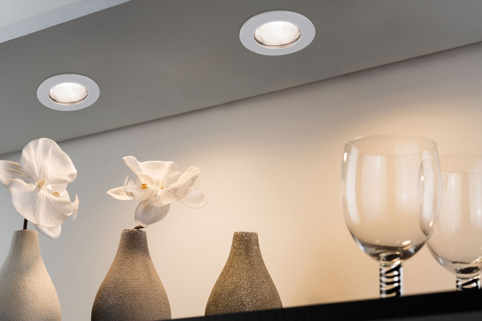 Zwei Einbaustrahler beleuchten Vasen mit weißen Orchideen und Weingläser.