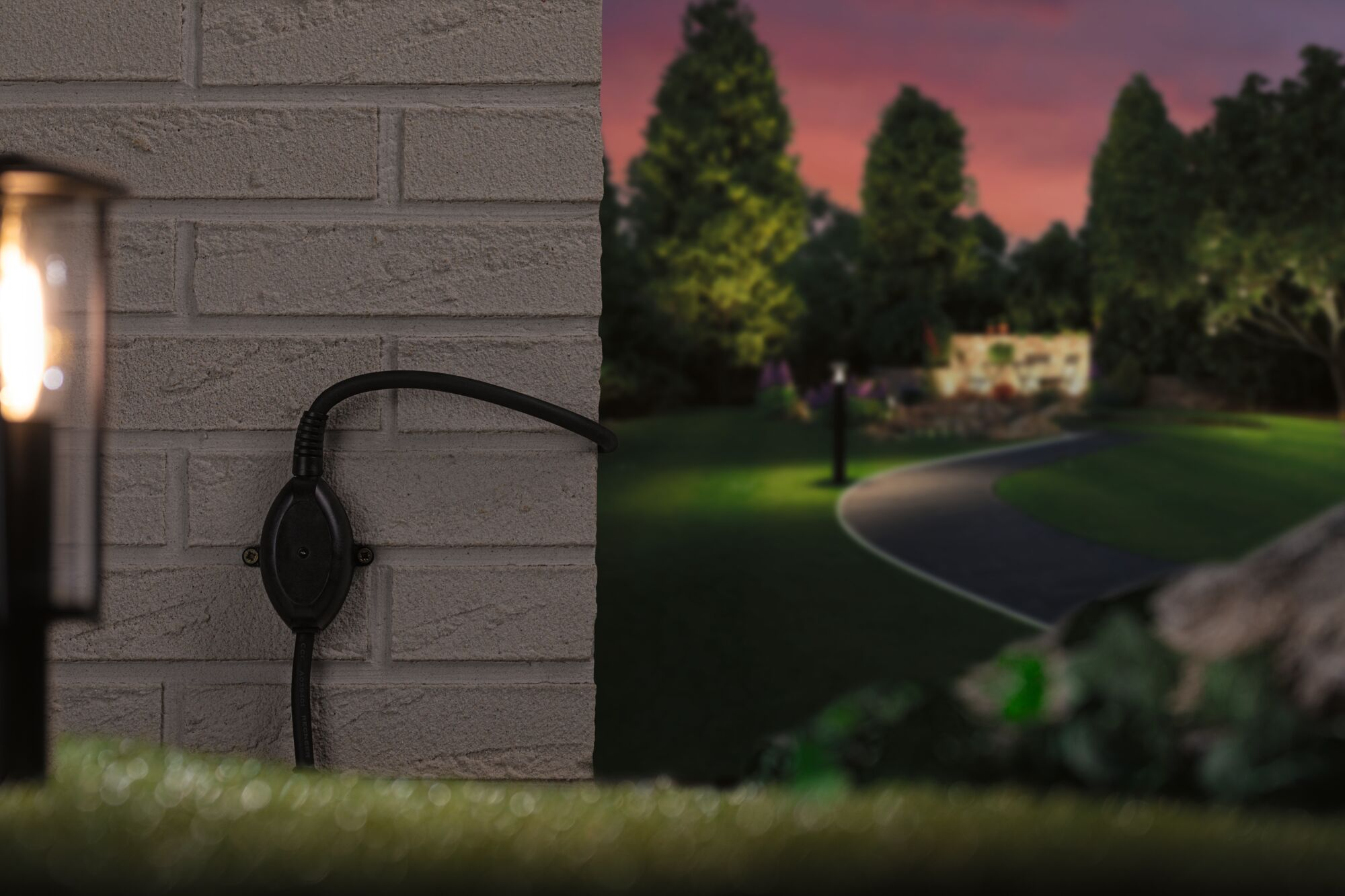 Ein schwarzes Kabel mit Stecker hängt an einer hellgrauen Ziegelwand neben einem Garten im Dämmerlicht.