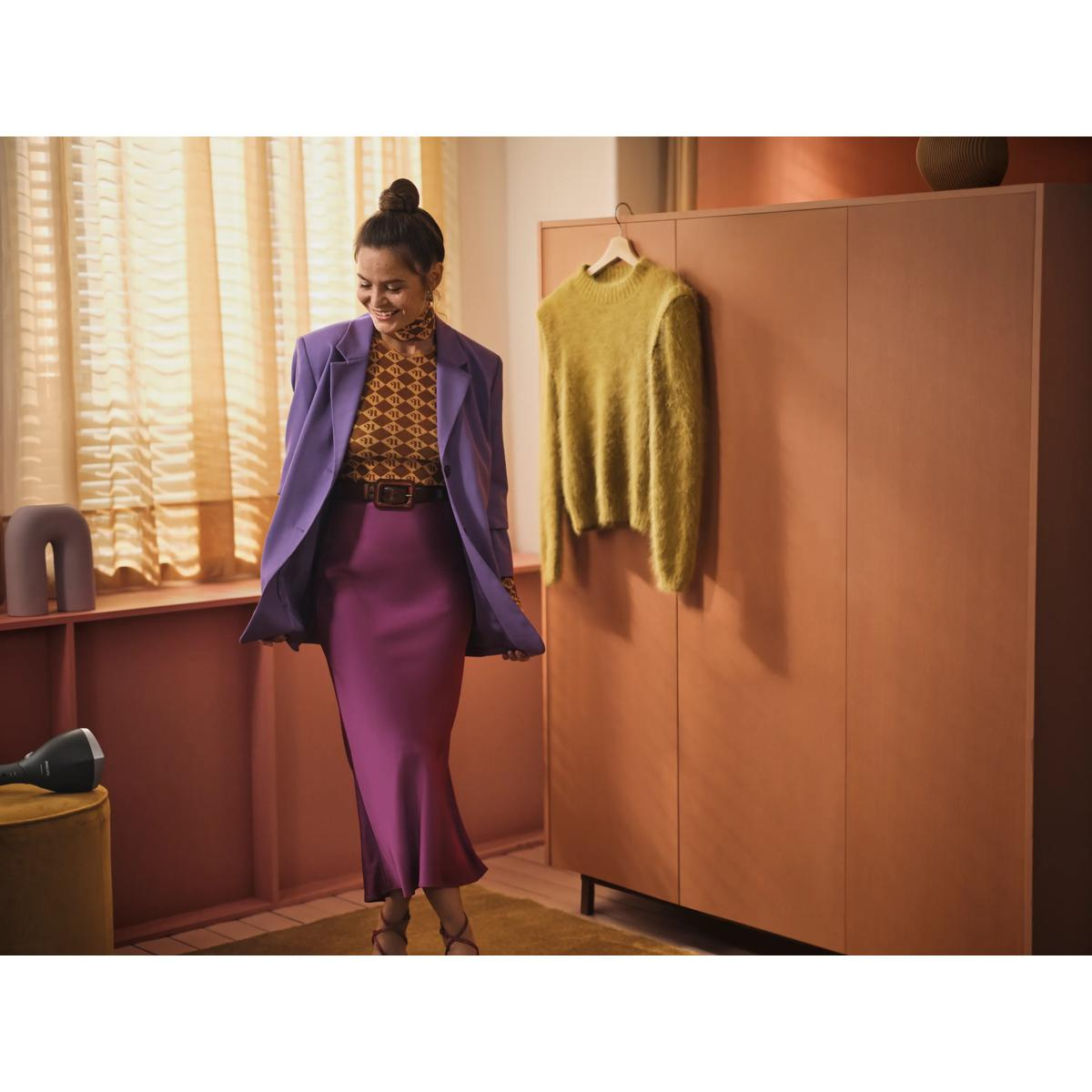 Donna in giacca viola e gonna prugna accanto ad armadio con maglione giallo.