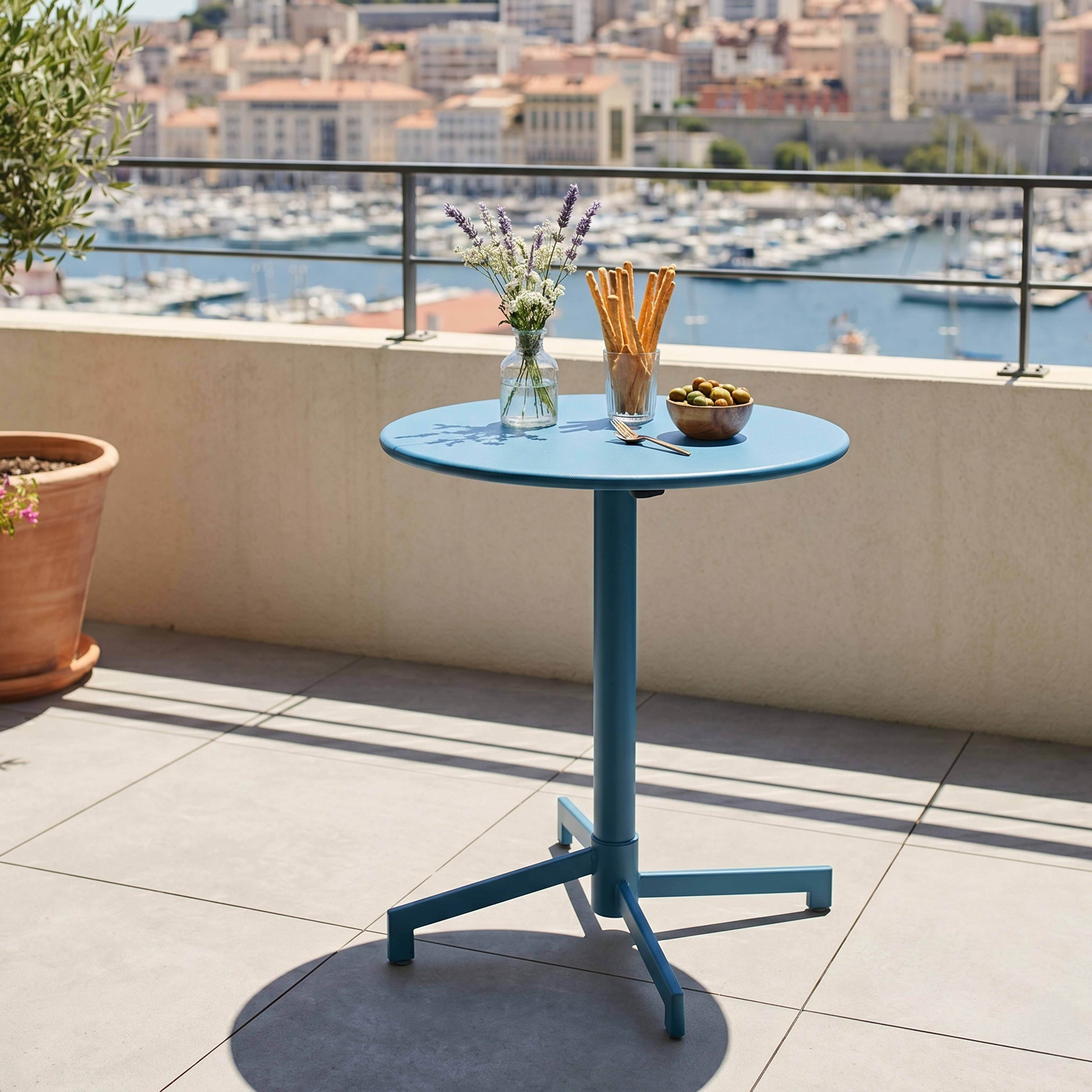Ein blauer Tisch mit Blumen, Grissini, Oliven und einer Gabel auf einer Terrasse mit Yachten im Hintergrund.