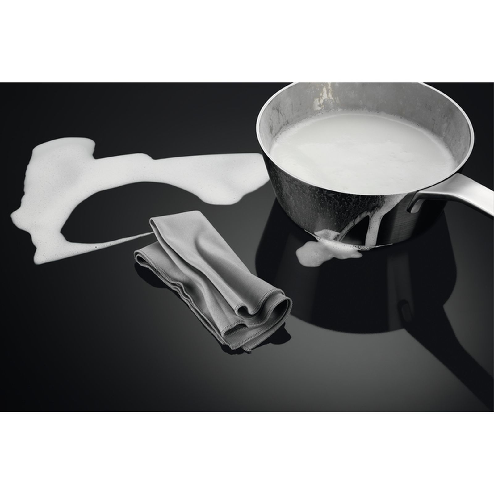 Pentola con liquido bianco su piano cottura nero, accanto a un panno grigio con schiuma.