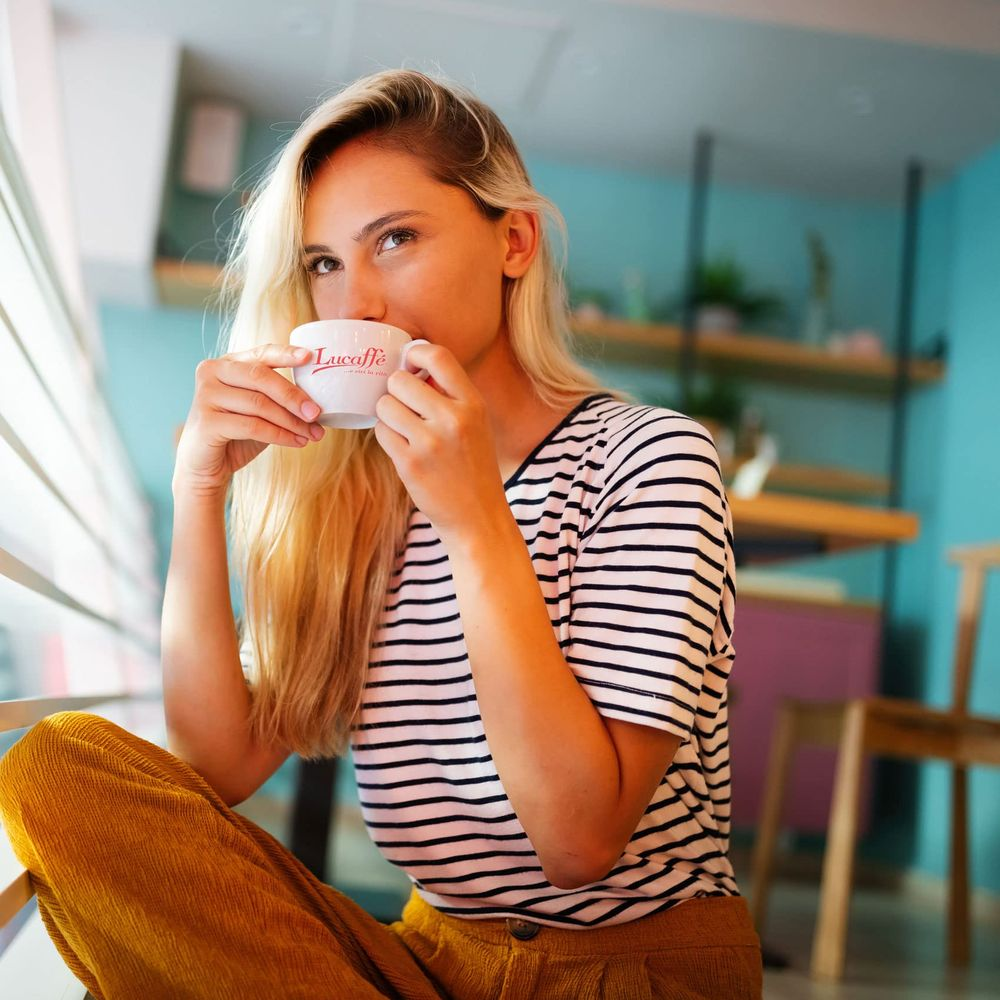 Eine Frau mit blonden Haaren trinkt aus einer weißen Tasse mit rotem Logo.