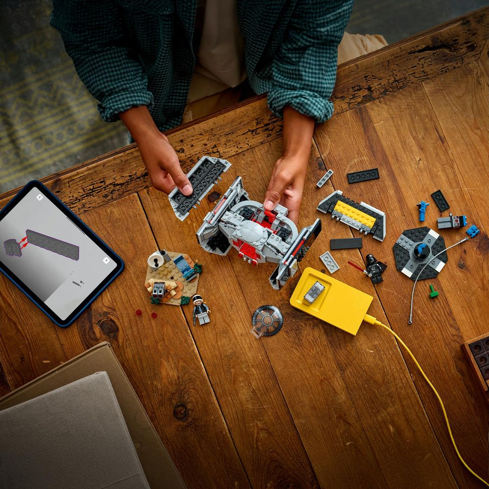 Mani che costruiscono un'astronave grigia e rossa con mattoncini Lego su un tavolo di legno. Il tablet mostra le istruzioni.