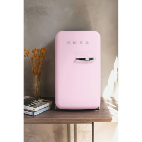 Un refrigerador rosa con asa cromada se sienta en un estante junto a libros y flores secas.