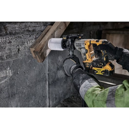 Una persona con guantes y chaqueta verde usa un taladro percutor DeWalt amarillo en pared de hormigón.
