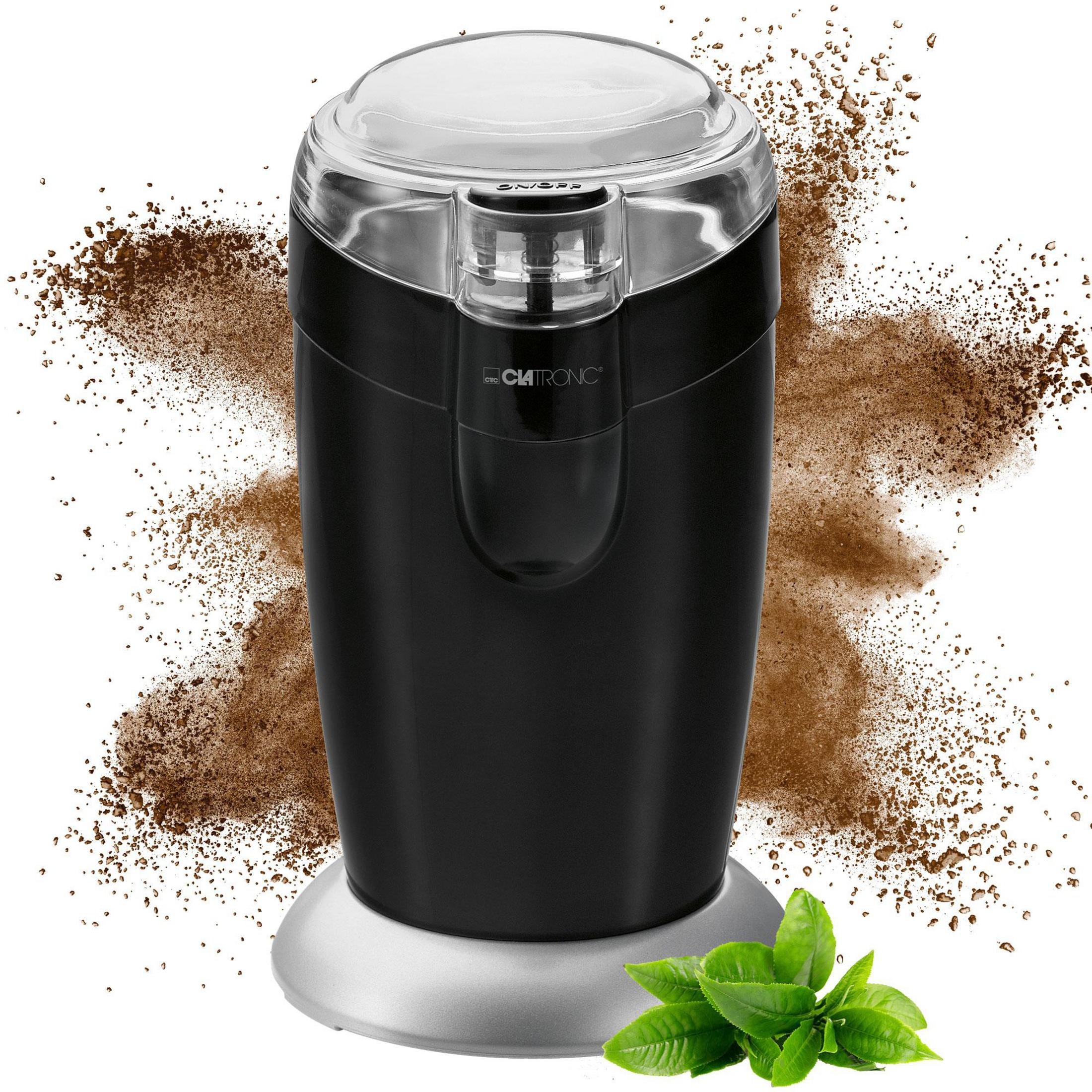 Tritacaffè nero con coperchio trasparente, circondato da fondi di caffè e foglie verdi.