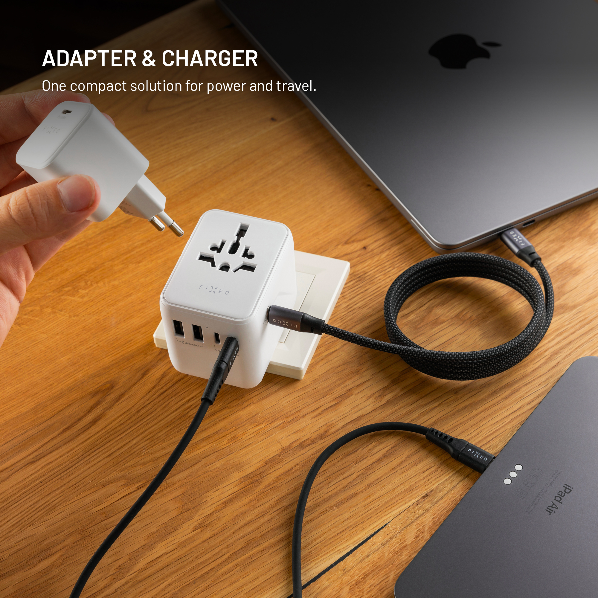 Ein weißer Reiseadapter mit USB-Anschlüssen steckt in einer Steckdose. Kabel verbinden ihn mit einem MacBook und iPad.