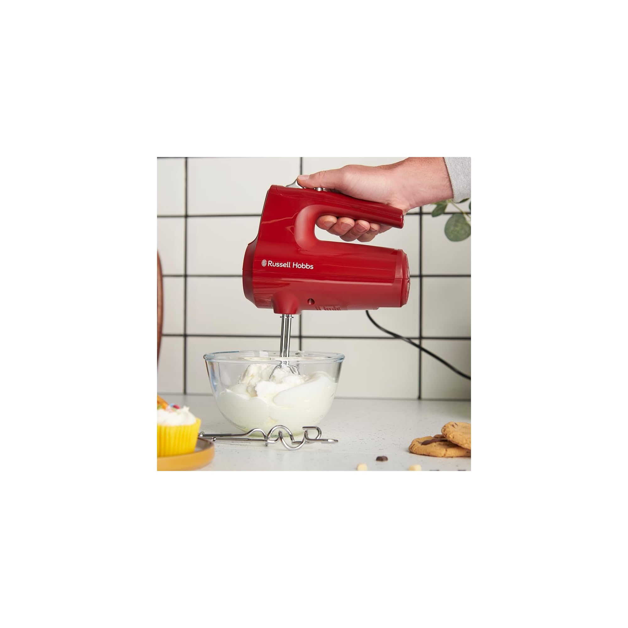 Rote Handrührgerät mixt weiße Creme in Glasschüssel neben Cupcake.