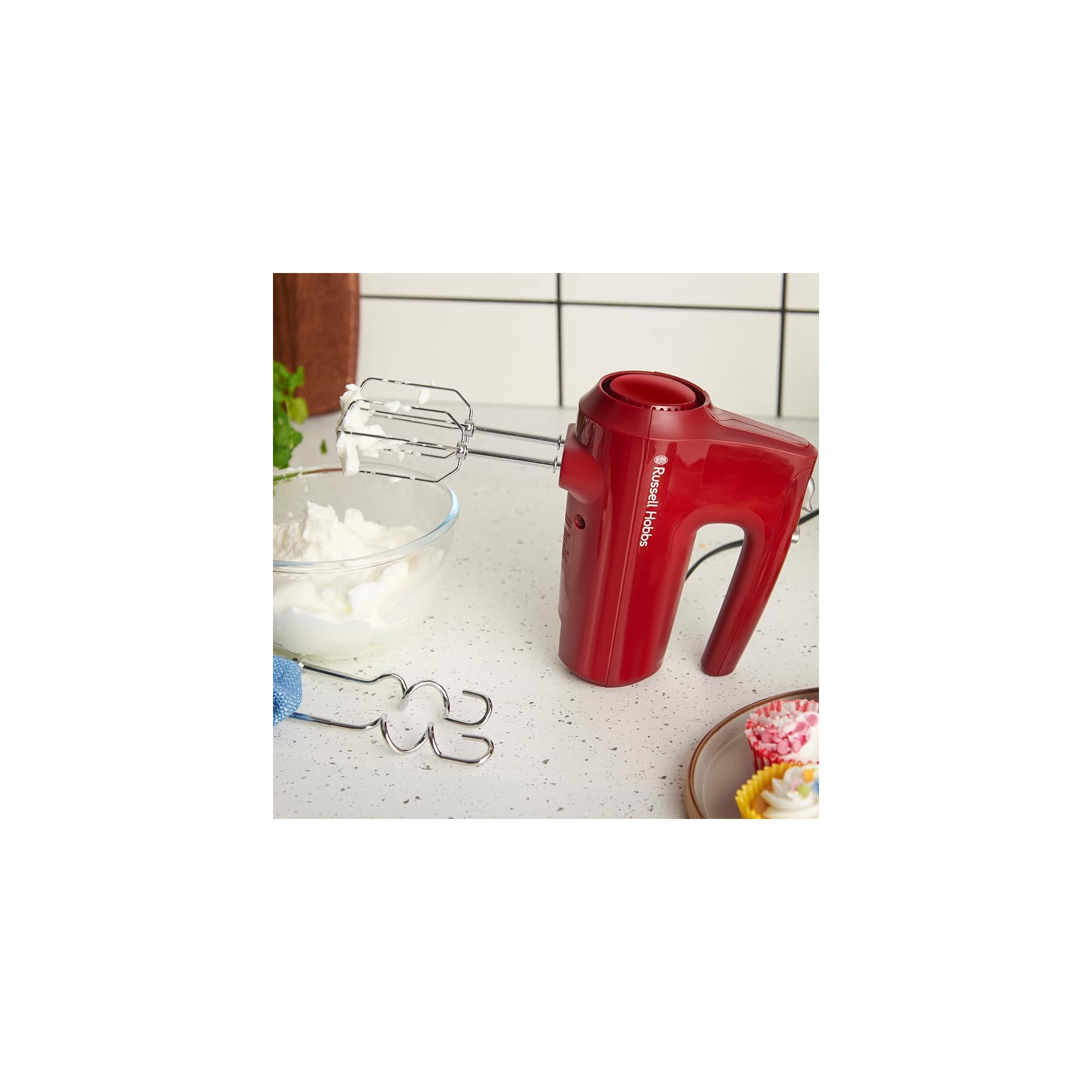 Ein roter Handmixer mit Schneebesen steht neben einer Schüssel mit Schlagsahne und Cupcakes.