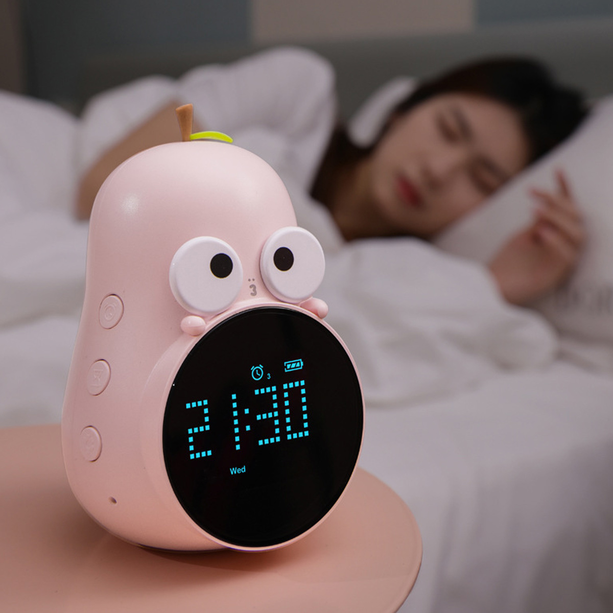 Ein rosafarbener Wecker in Form einer Frucht mit großen Augen schläft neben einer Frau im Bett.