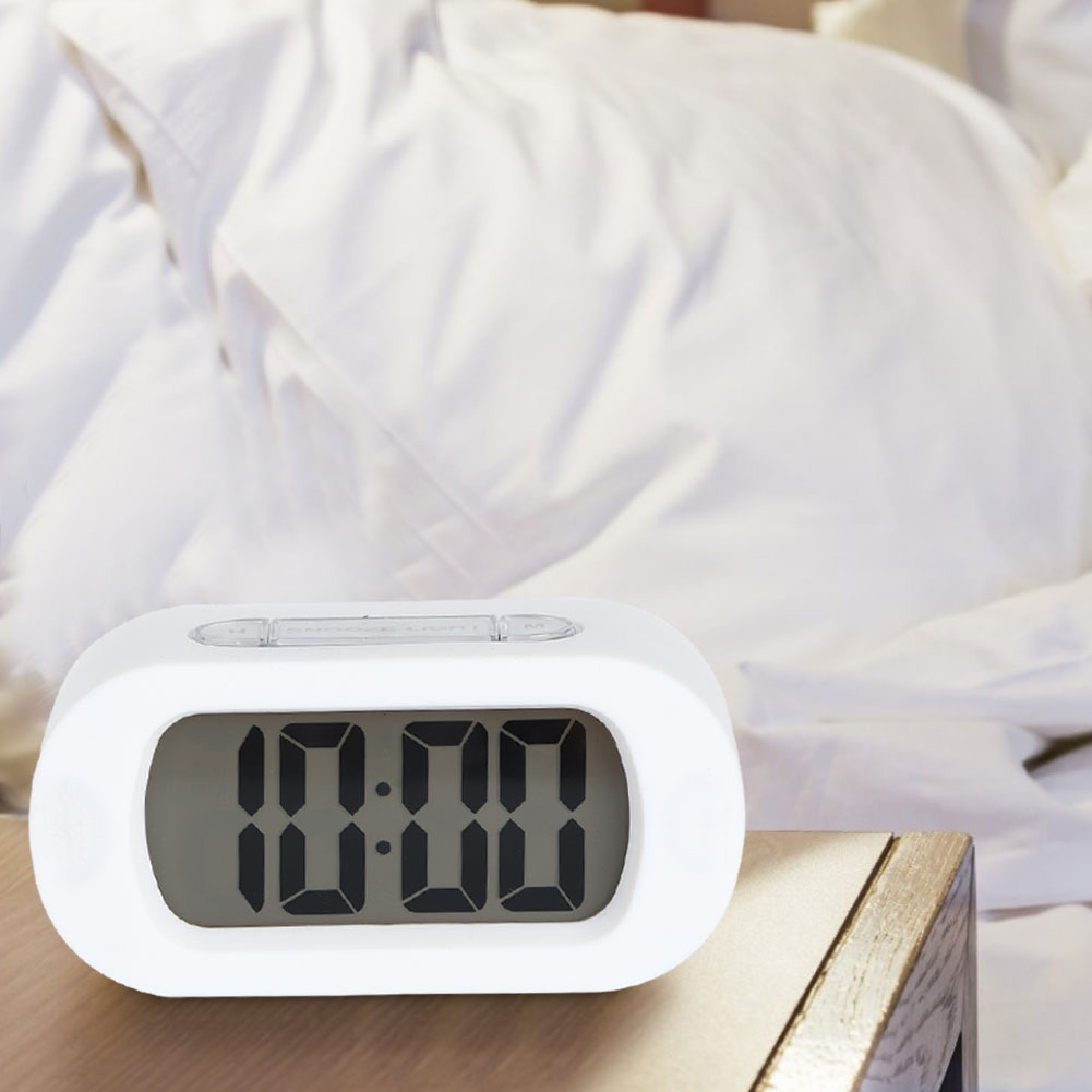 Ein weißer digitaler Wecker zeigt 10:00 Uhr auf einem Nachttisch neben einem Bett.