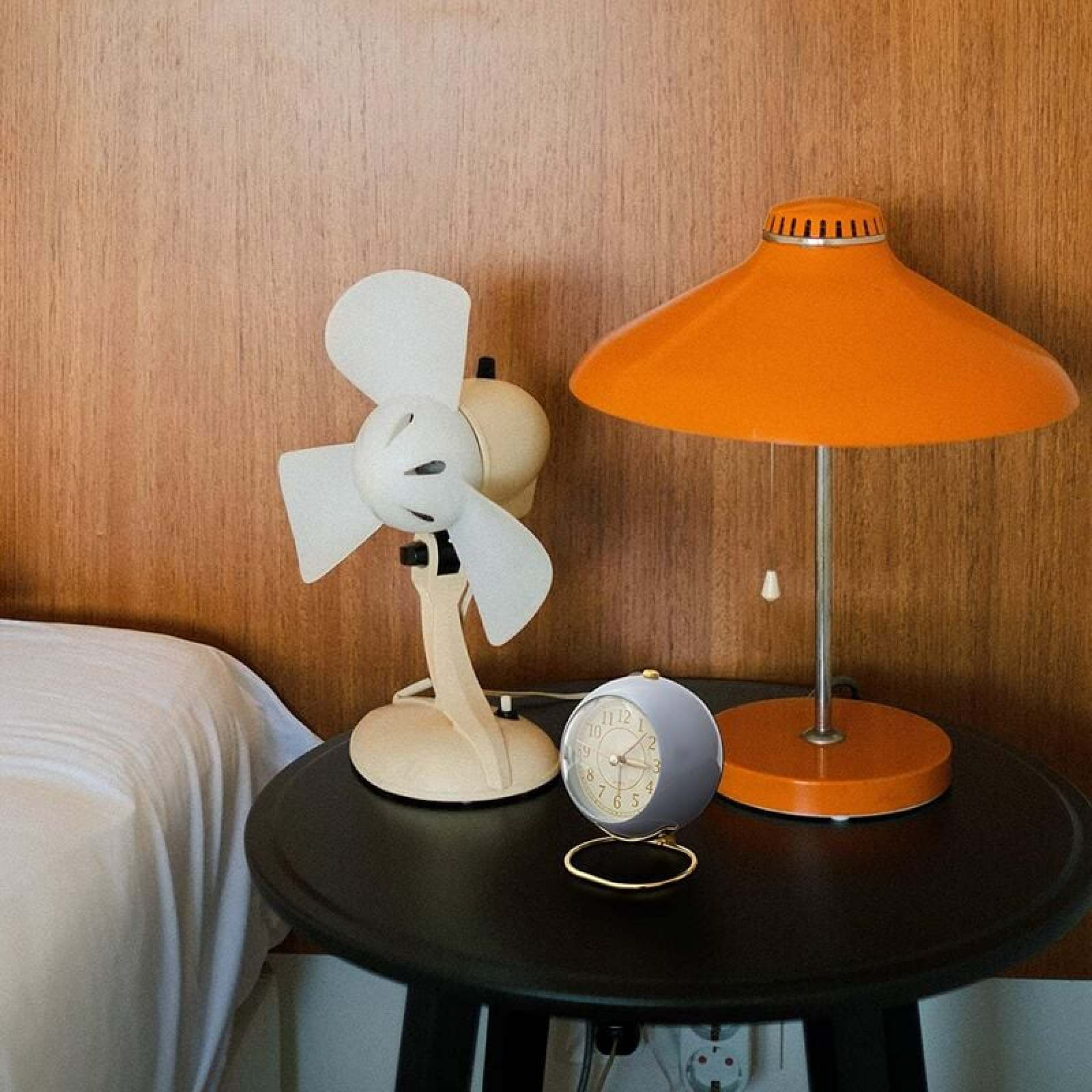 Ein Nachttisch mit Ventilator, Lampe und Wecker vor einer Holzvertäfelung.