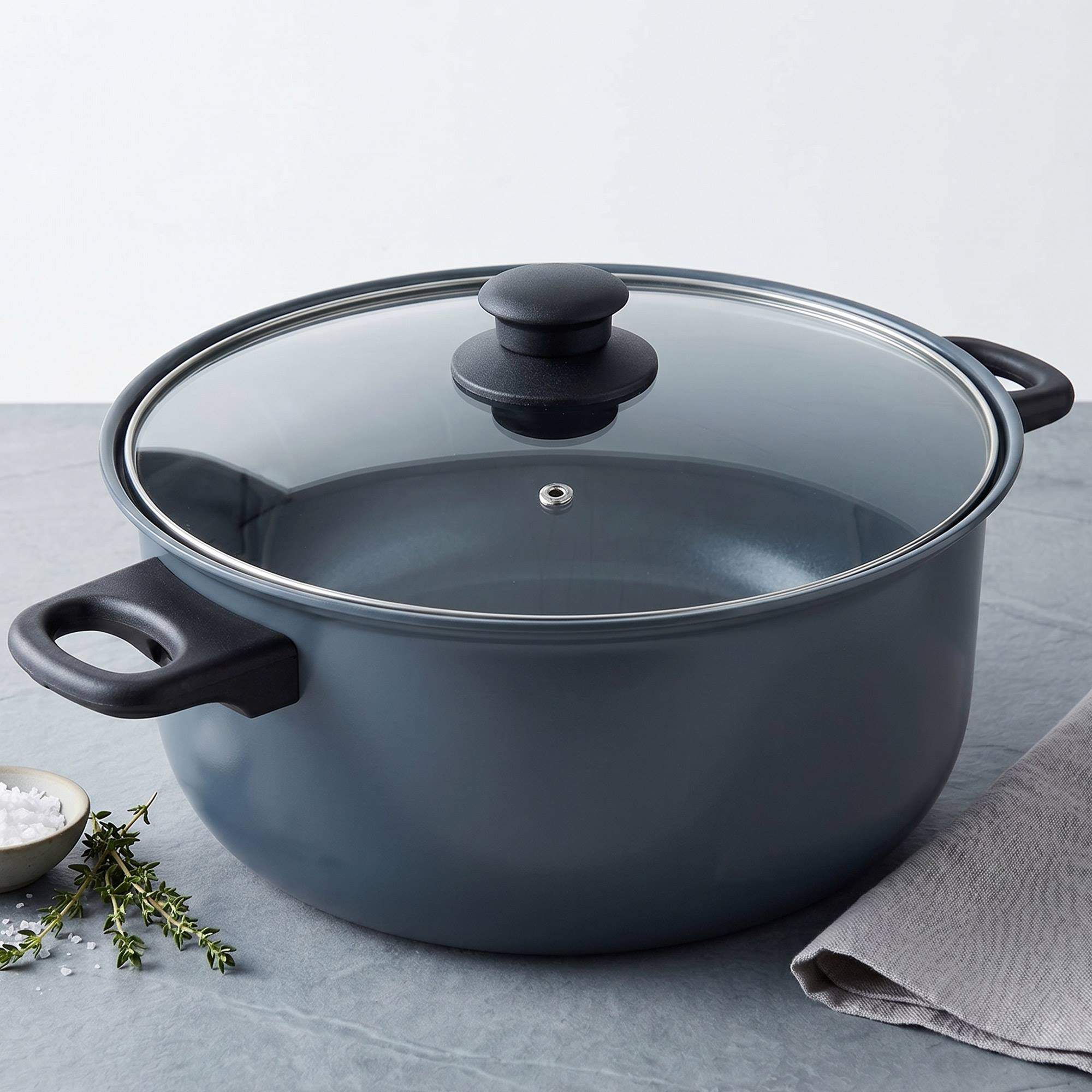 Una olla oscura con tapa de cristal y asas negras sobre una superficie gris.