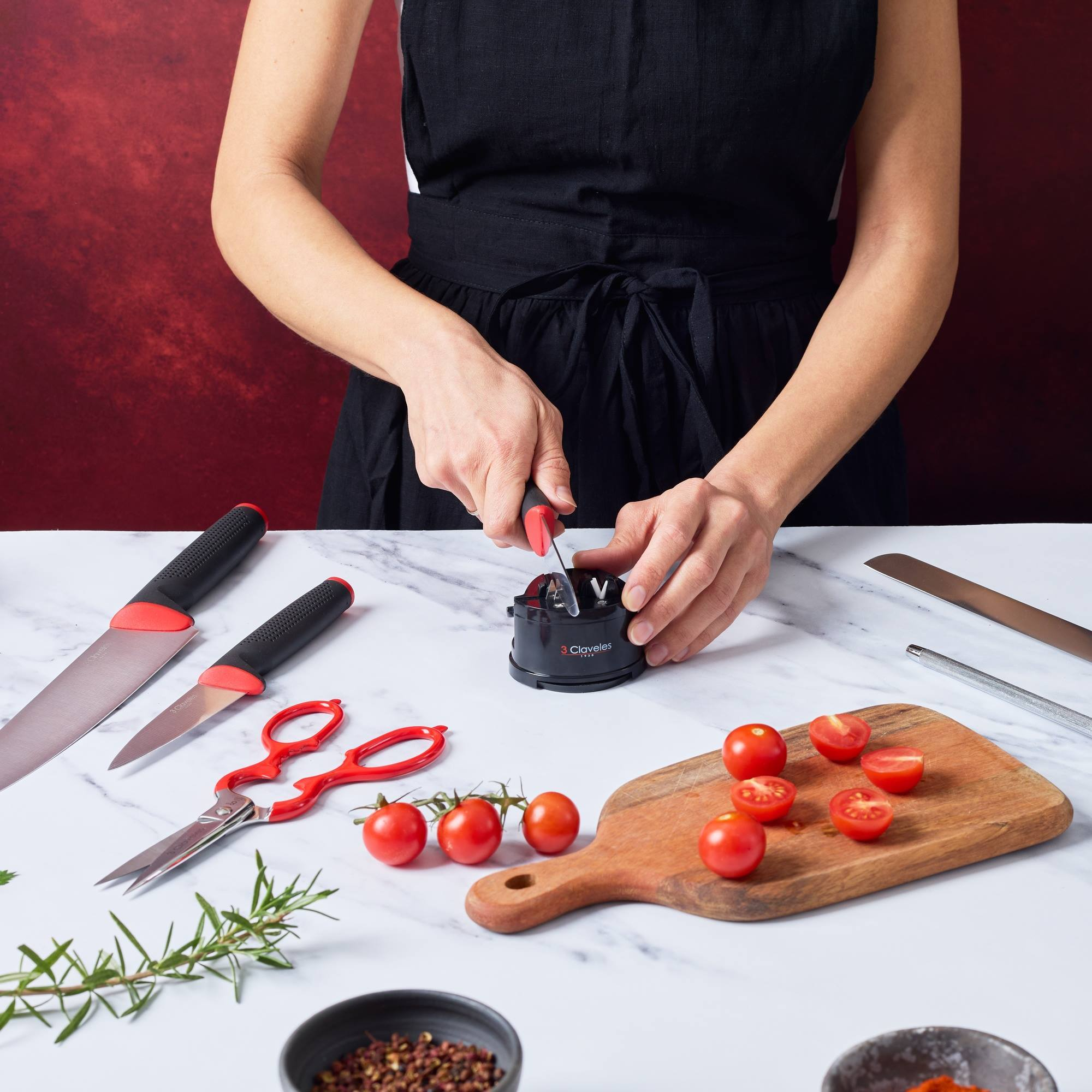 Persona afila cuchillo con afilador negro. Tomates, hierbas, tijeras, cuchillos en mesa mármol.