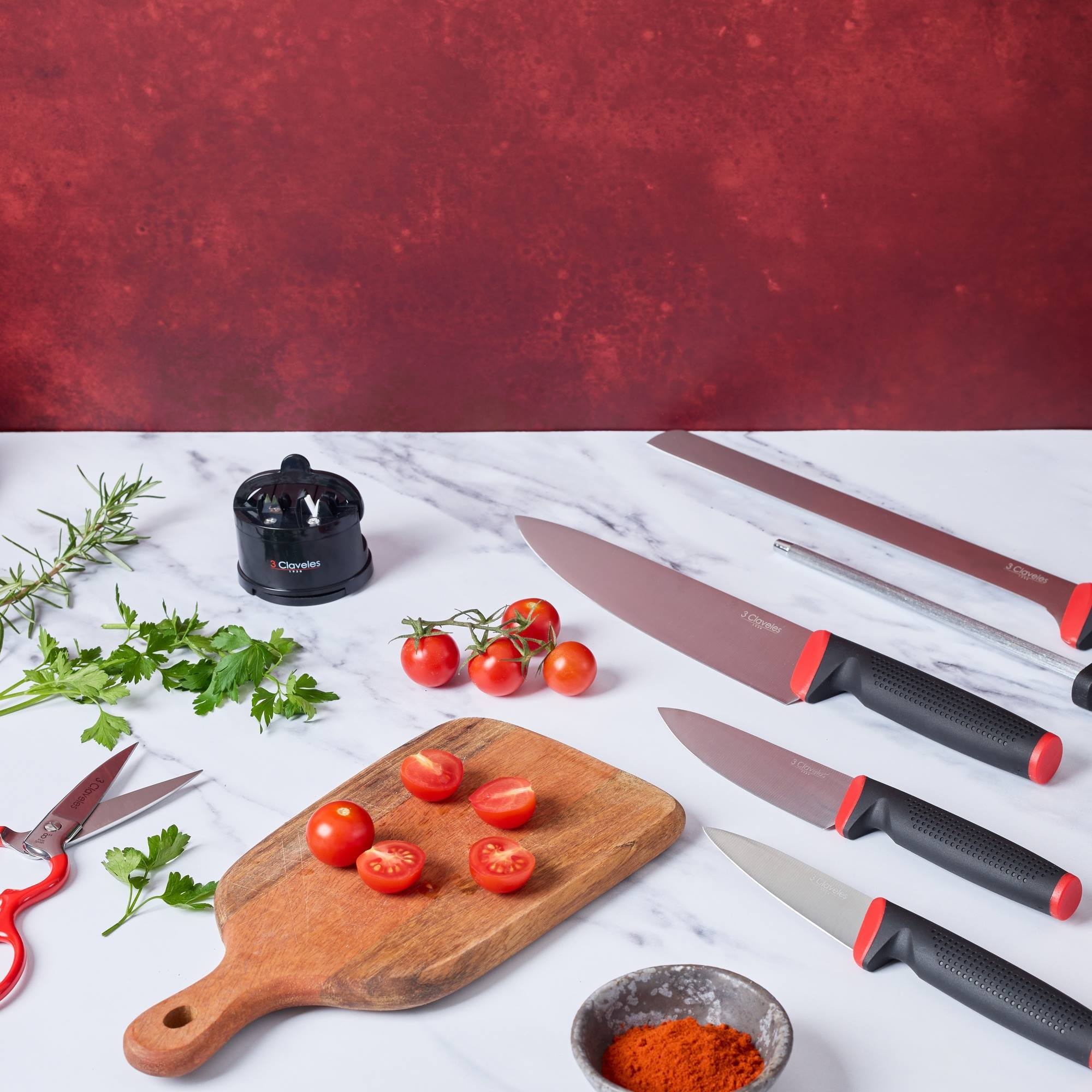Cuchillos, tomates y hierbas sobre superficie de mármol con fondo rojo.
