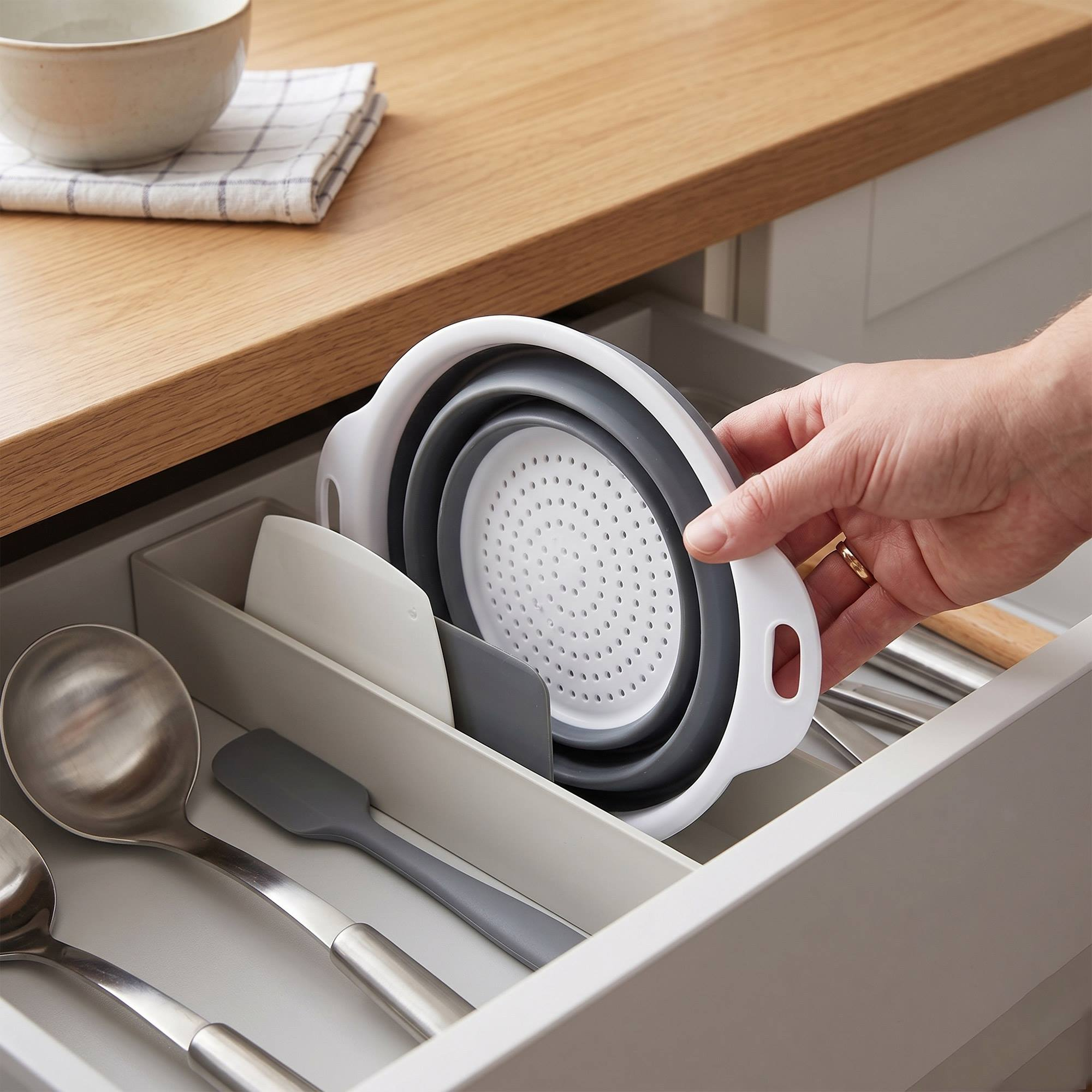 Una mano coloca un colador plegable en un cajón de cocina con utensilios.