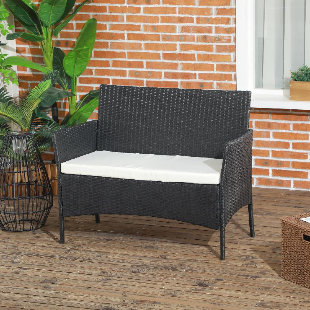 Un divanetto in vimini nero con cuscino bianco siede su un ponte di legno con piante e muro.
