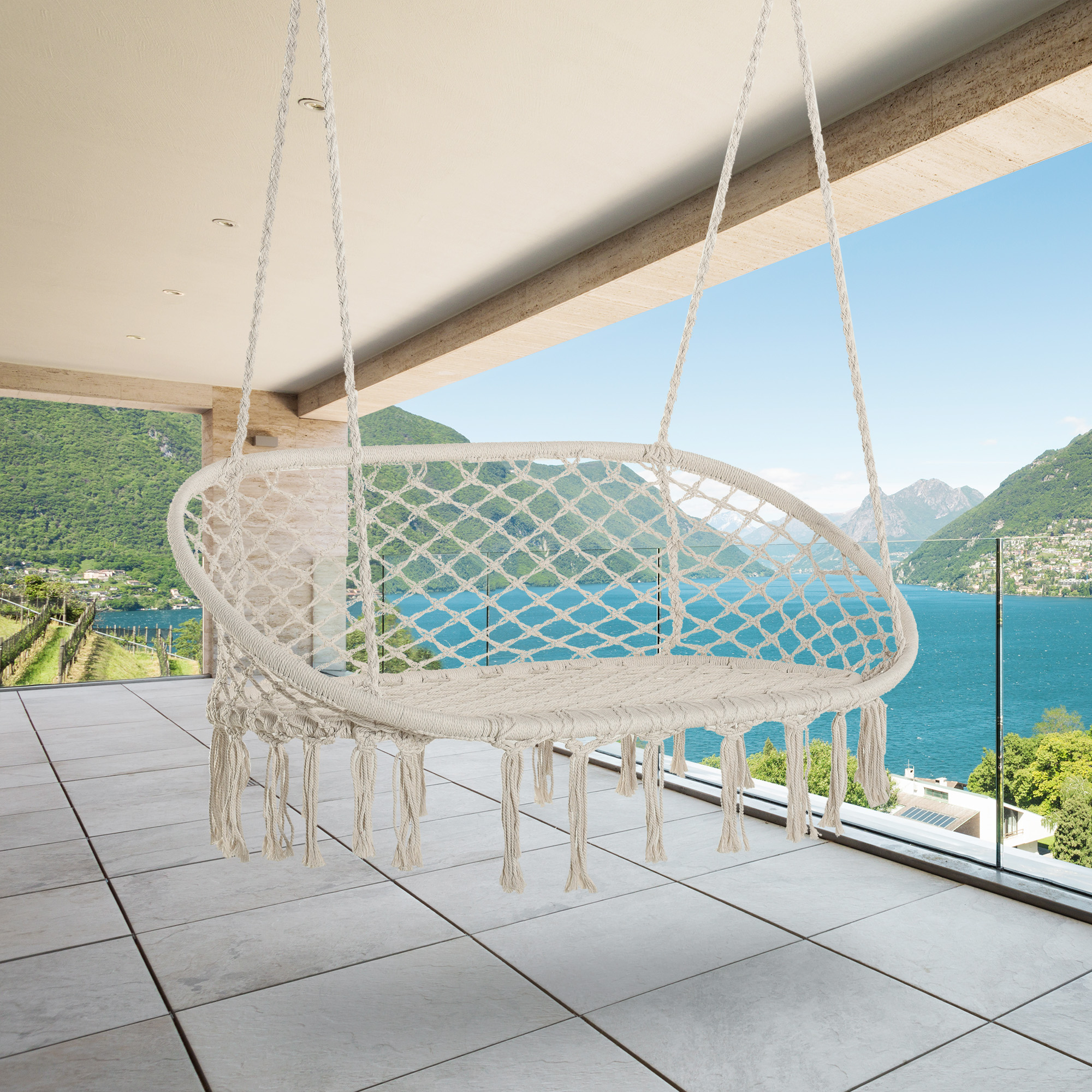 Una sedia amaca in macramè è appesa su un balcone con vista su un lago e montagne.