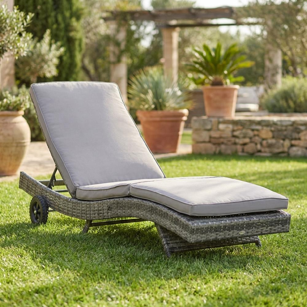 Una chaise longue in rattan grigio con cuscini riposa sull'erba verde in un giardino.