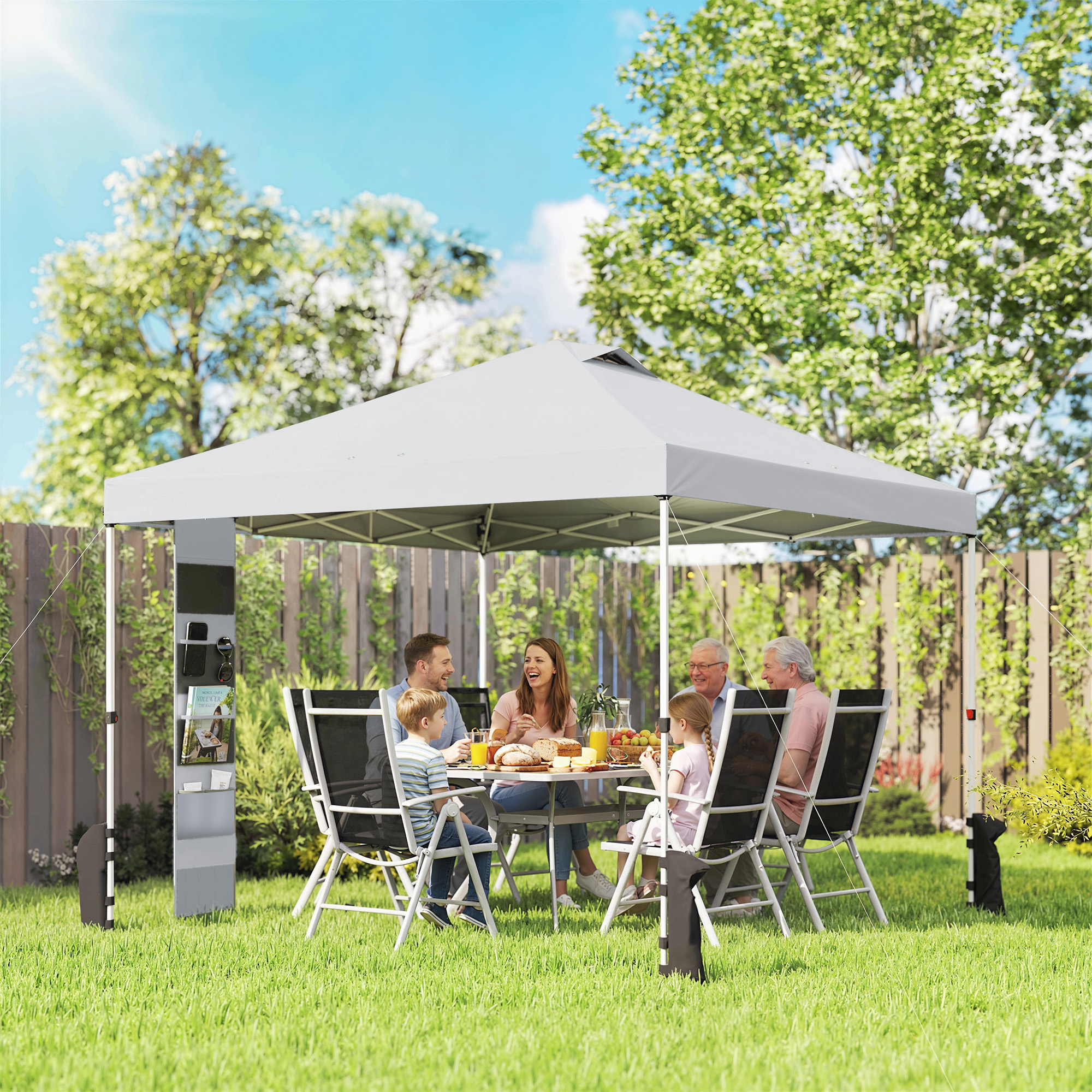 Una famiglia cena sotto un gazebo in una giornata di sole, con alberi e una recinzione.