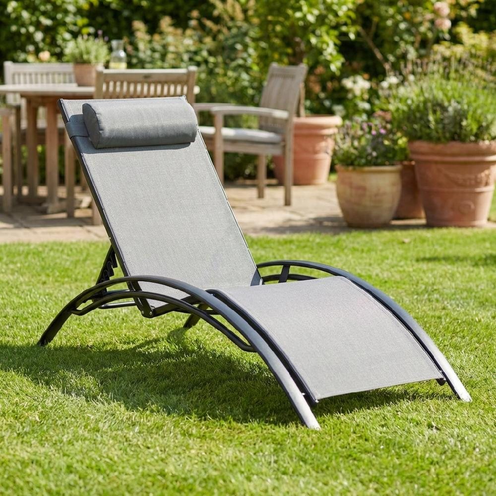 Sdraio grigio con cuscino sull'erba, mobili da giardino sullo sfondo.
