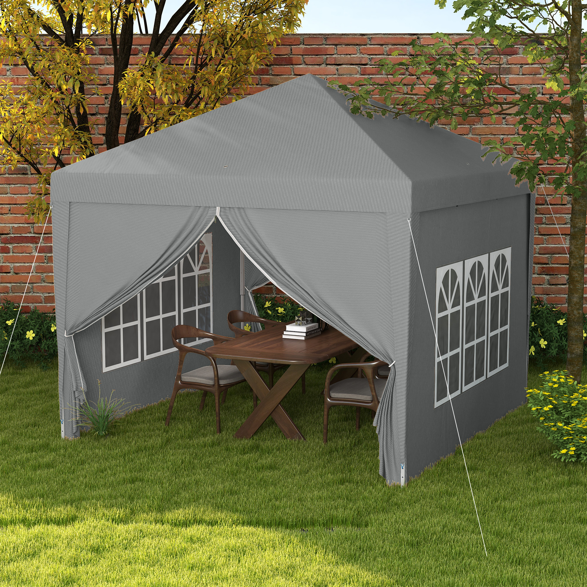 Un gazebo grigio con tavolo e sedie è allestito su un prato verde, con un muro di mattoni e alberi sullo sfondo.