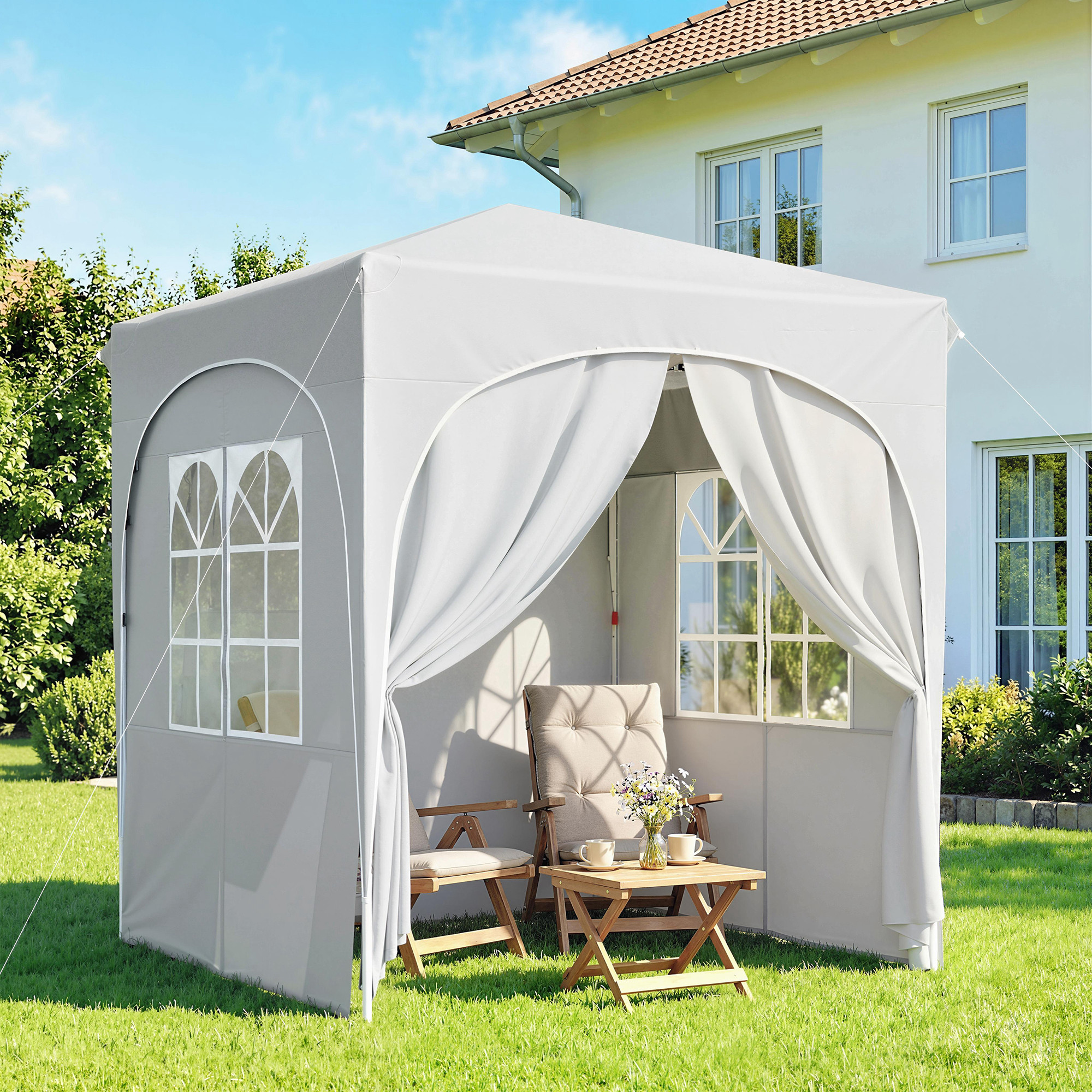Un gazebo grigio con finestre, due sedie e un tavolino con tazze e fiori in giardino.