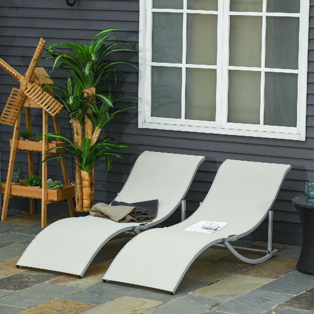 Due lettini prendisole di colore chiaro su un patio di pietra di fronte a una casa di legno scuro con finestra.
