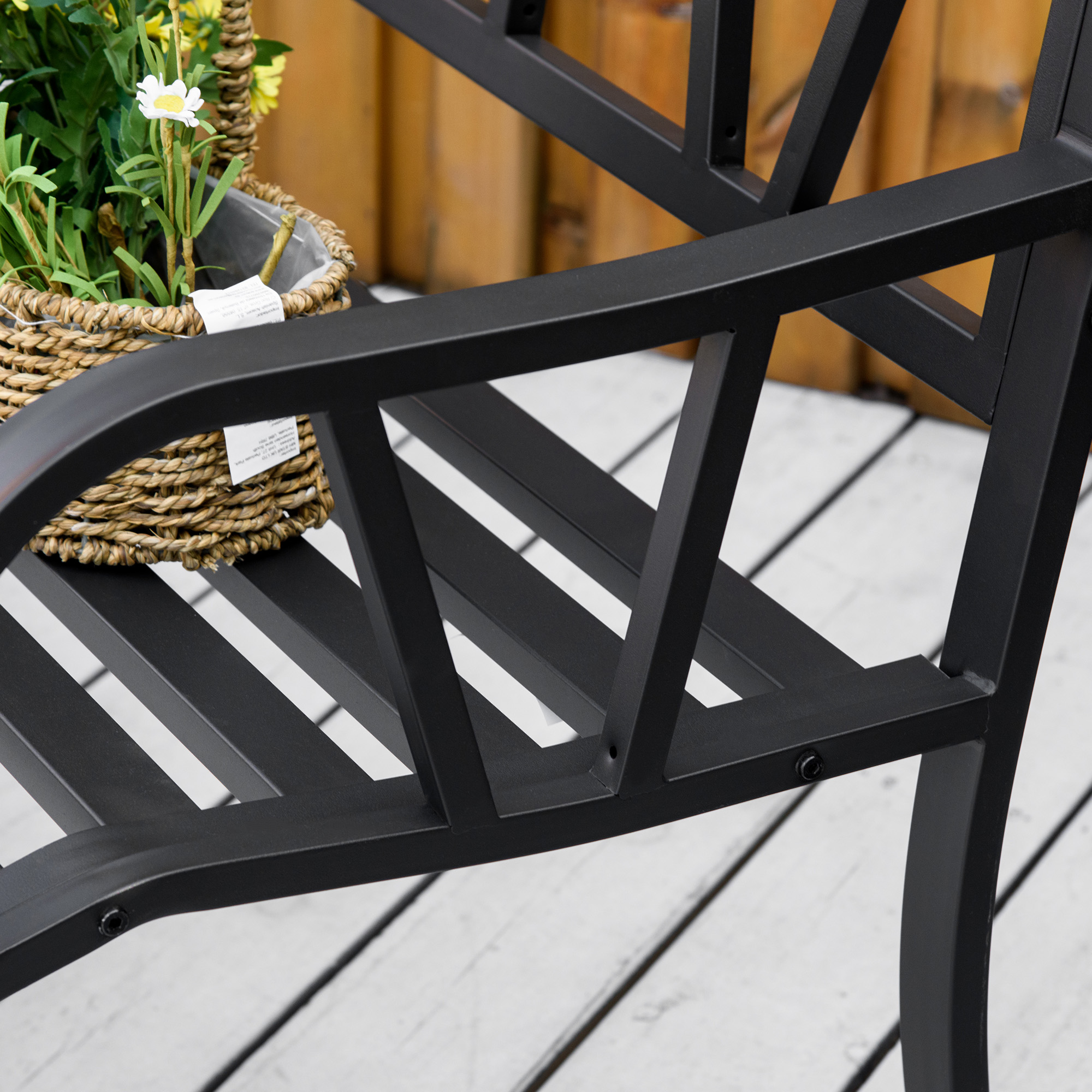 Una panchina in metallo nero con un cesto intrecciato contenente fiori. Sfondo in legno.