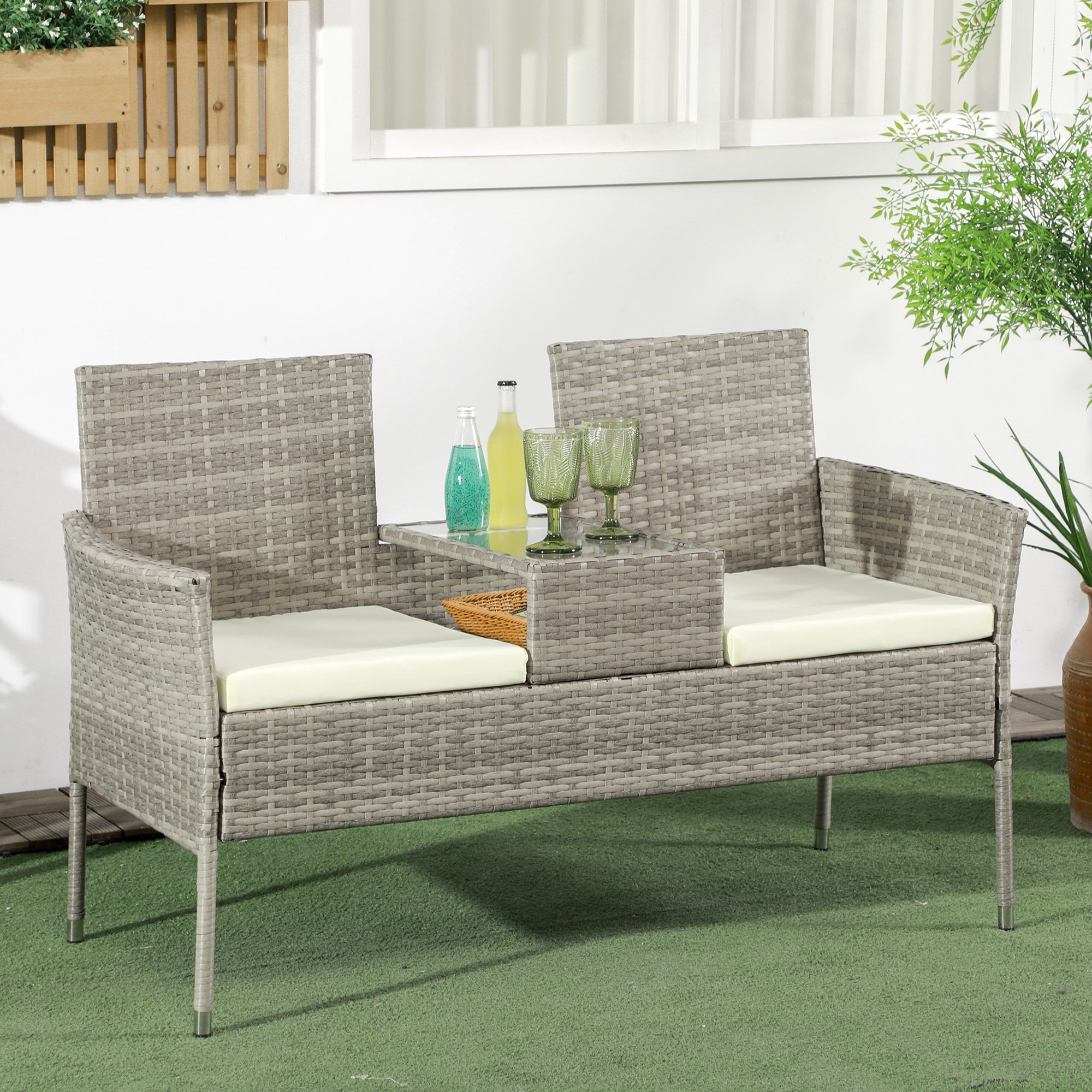 Divanetto in rattan grigio con cuscini beige chiaro, piano in vetro, bottiglie e bicchieri.
