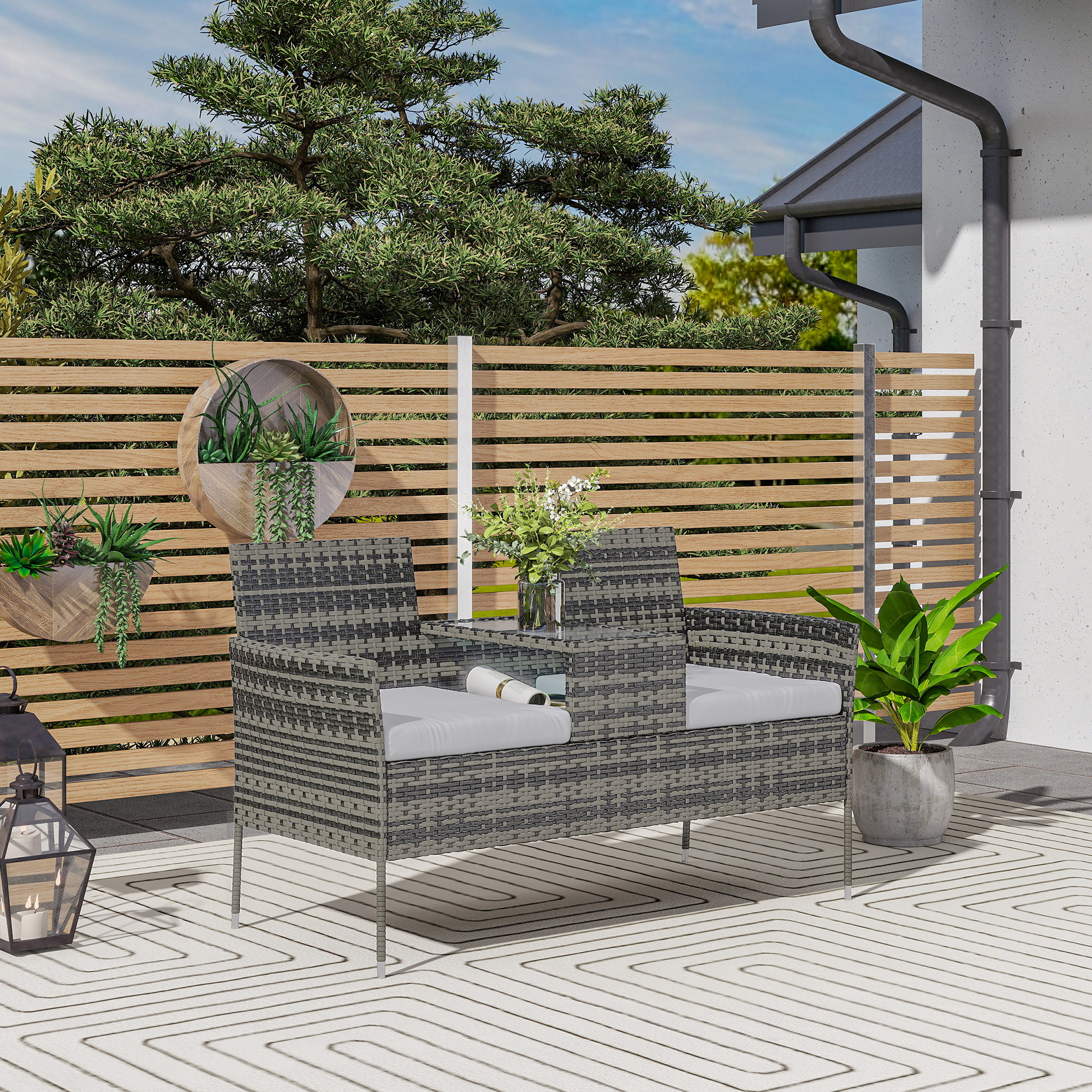 Divano in rattan grigio con cuscini bianchi su un patio con recinzione in legno e piante.