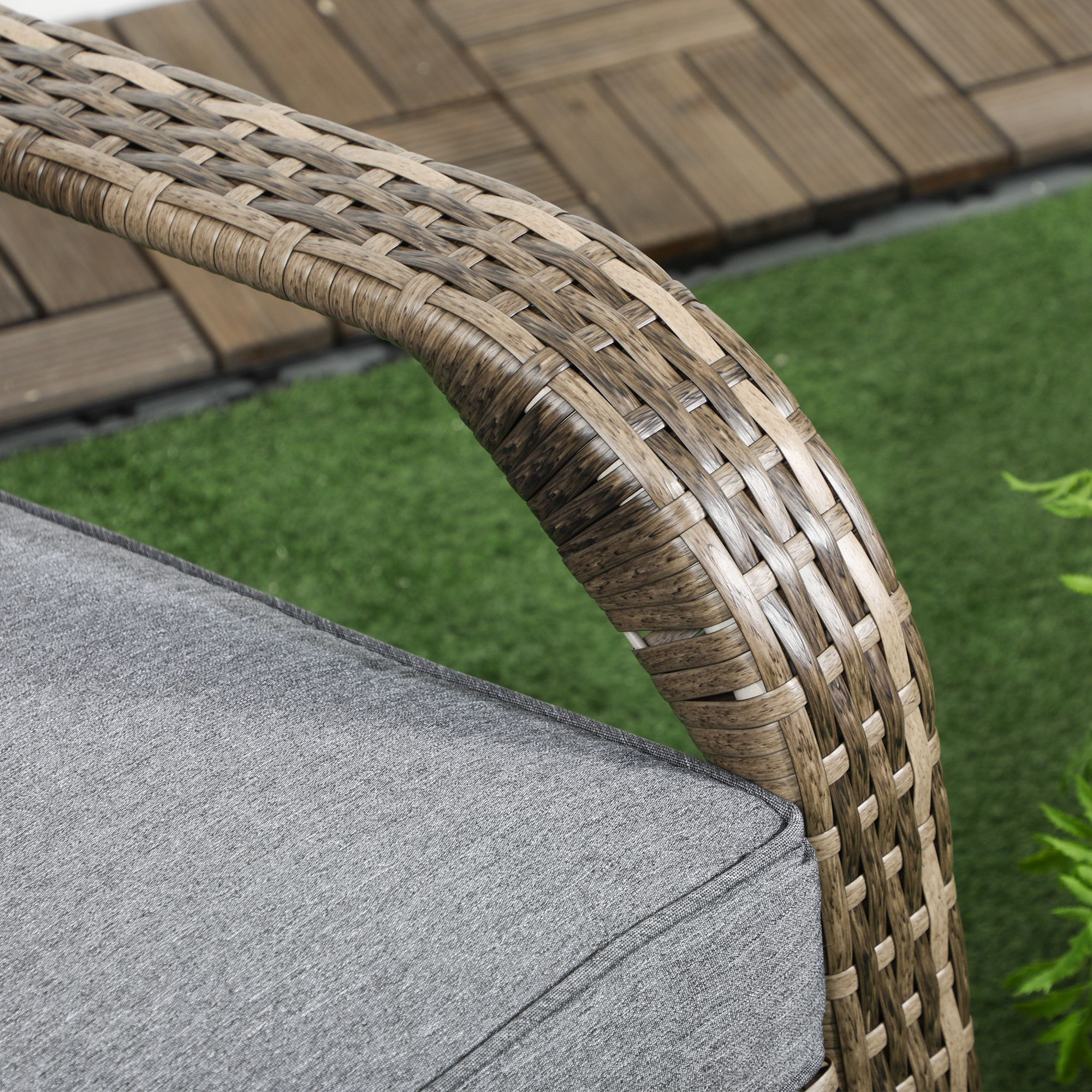 Bracciolo sedia in rattan marrone con cuscino grigio su erba sintetica e piastrelle di legno.